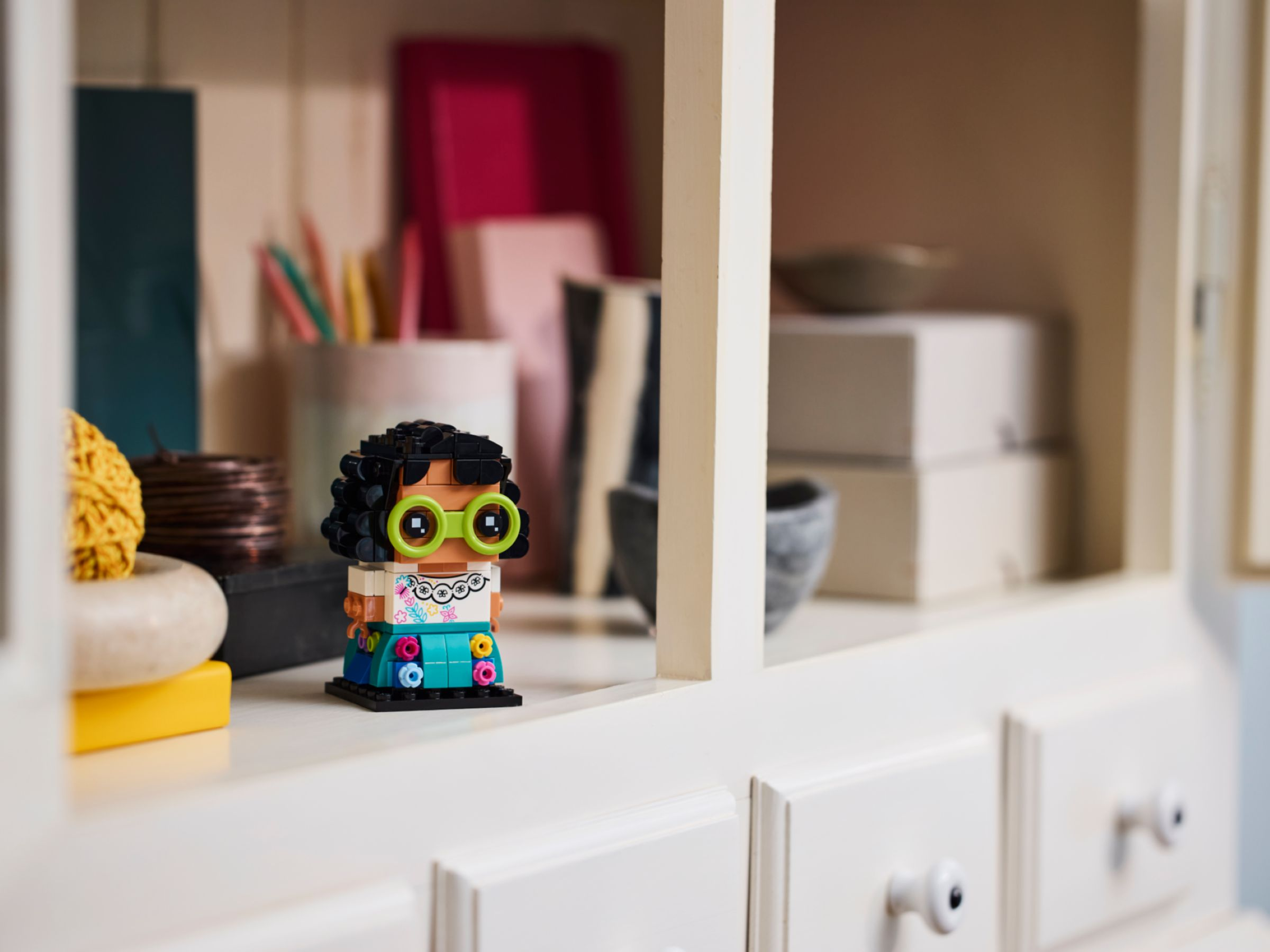 Lego-Figur einer Person mit Brille, schwarzem Haar und Blumenkleid. Sie steht auf einem Regal mit Gegenständen.
