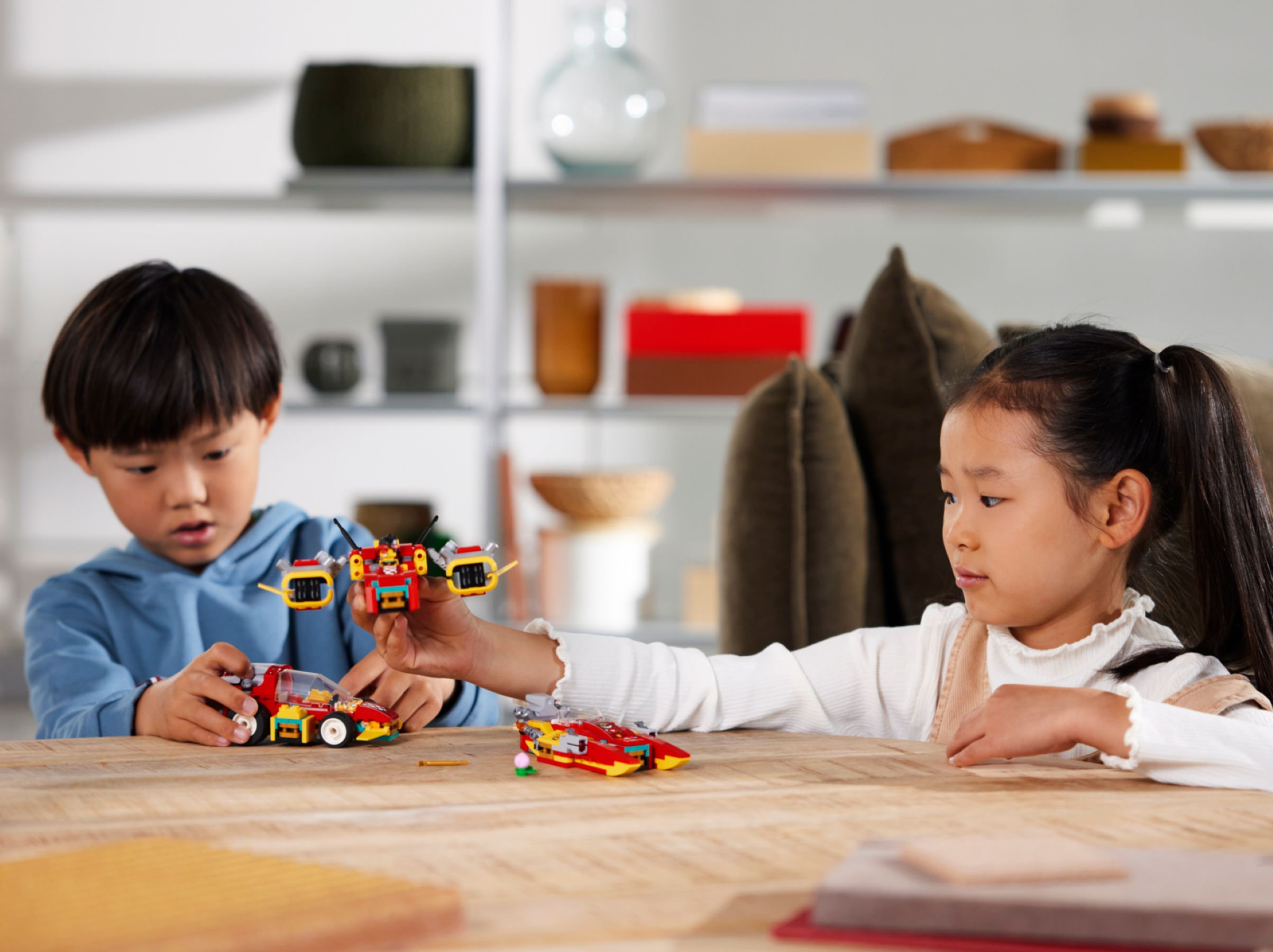 Zwei Kinder spielen mit Lego-Spielzeugen auf einem Holztisch. Ein Junge hält ein rotes Auto, ein Mädchen ein rotes Flugzeug.