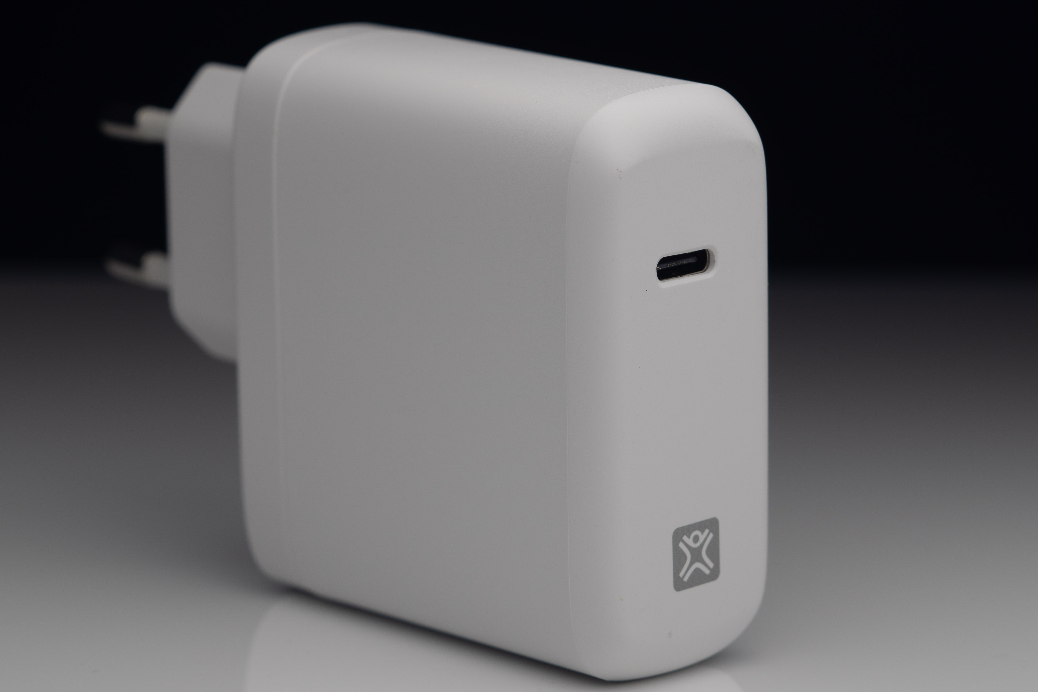 Un caricatore USB-C bianco con spina e logo. Su superficie riflettente, sfondo scuro.