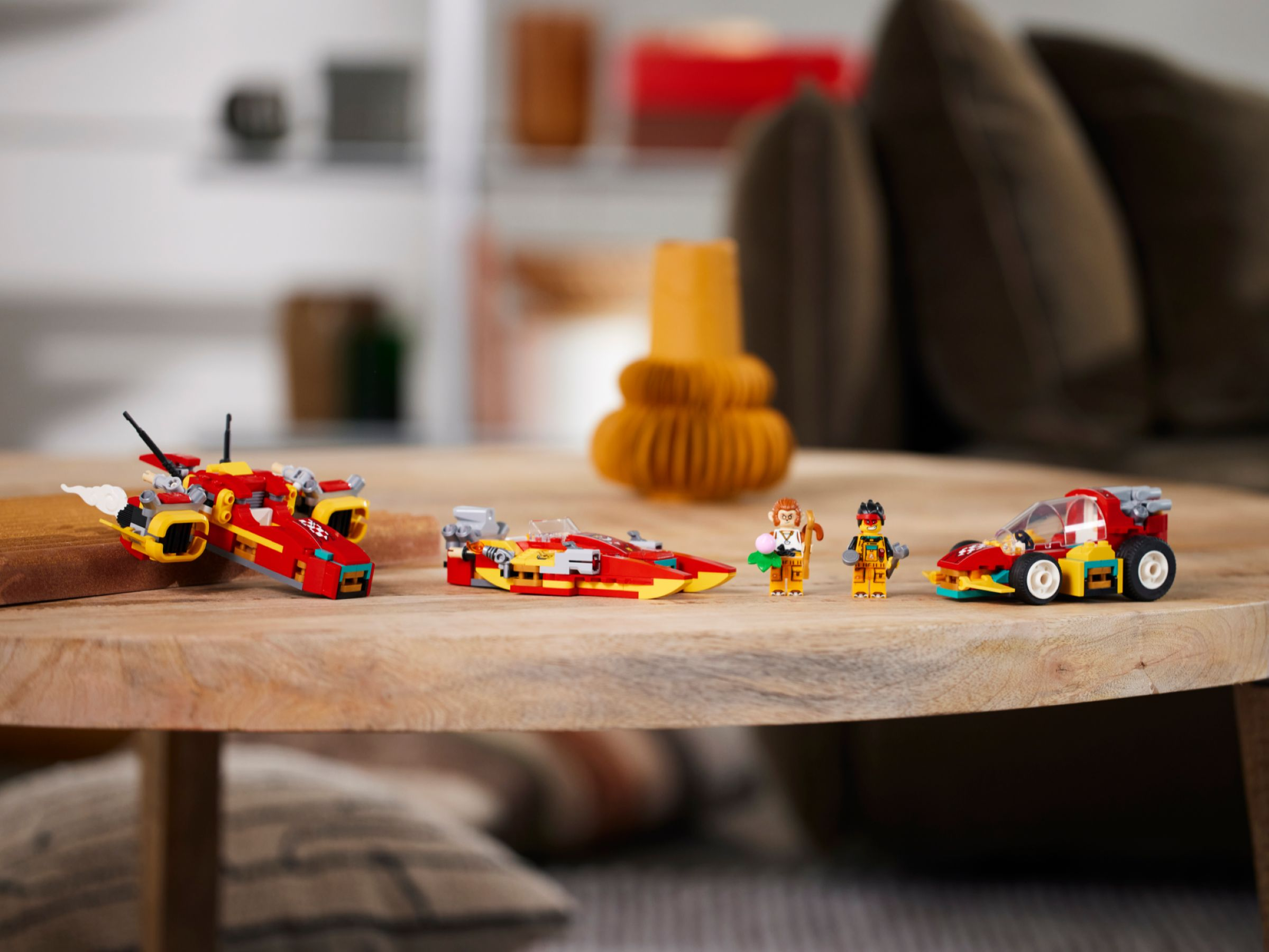 Auf einem Holztisch stehen LEGO-Sets: ein rotes Flugzeug, ein Boot, ein Auto und zwei Minifiguren. Der Hintergrund ist verschwommen.