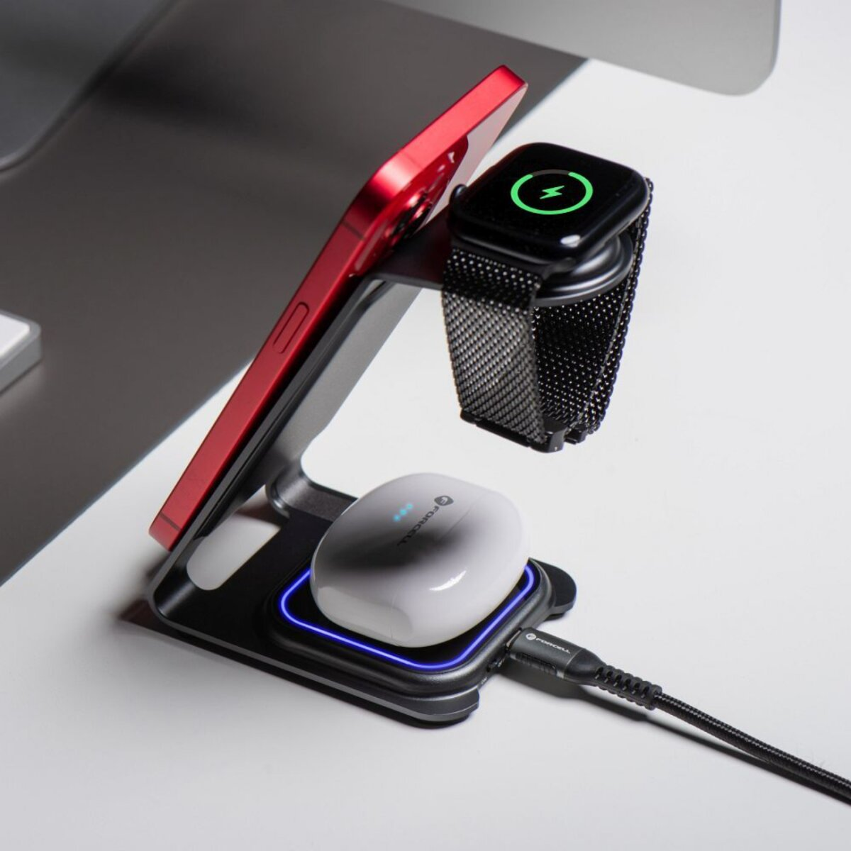 Eine Ladestation hält ein rotes Telefon, eine schwarze Uhr und weiße Ohrhörer. Alle werden geladen. Ein Kabel ist angeschlossen.