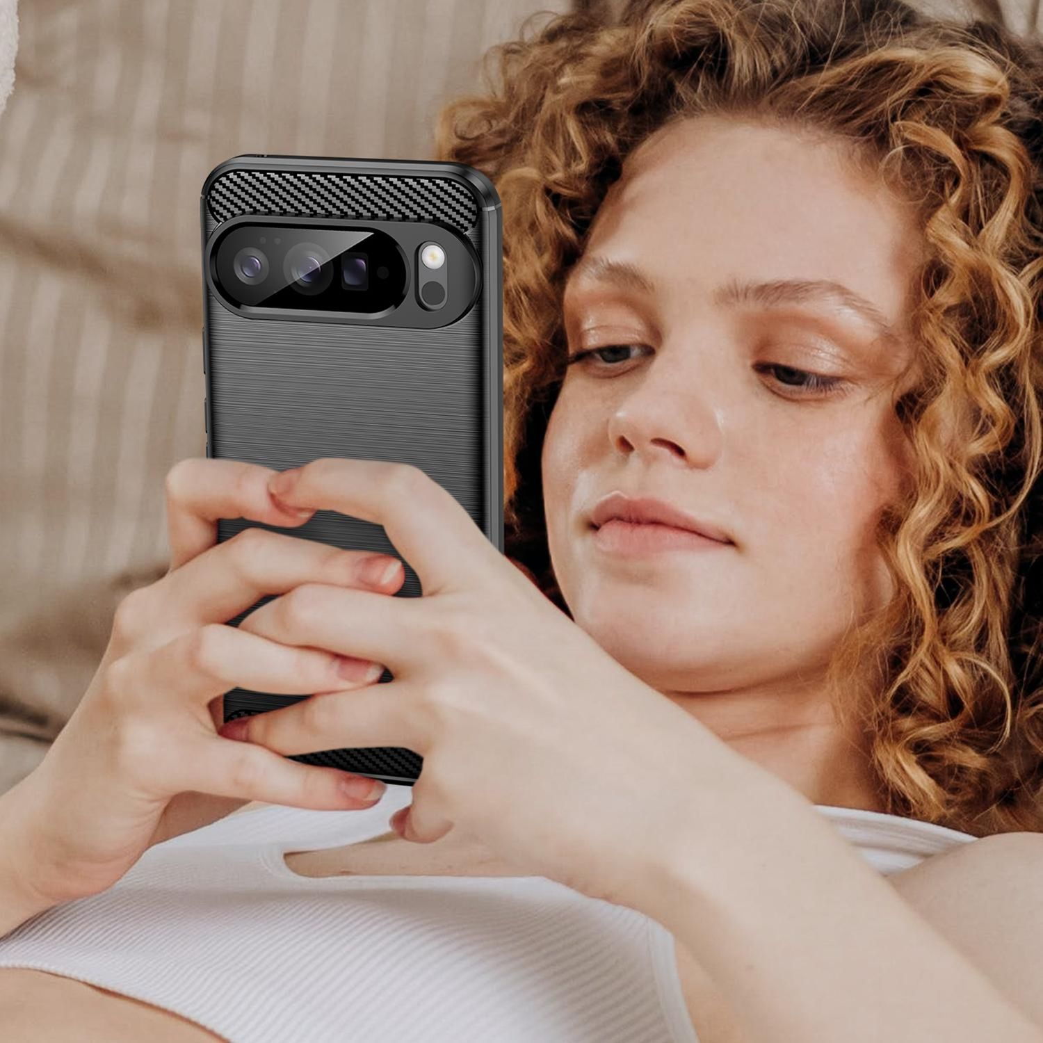 Eine Frau mit lockigem Haar hält ein schwarzes Telefon. Sie schaut auf den Bildschirm, mit einem weißen Oberteil, drinnen.