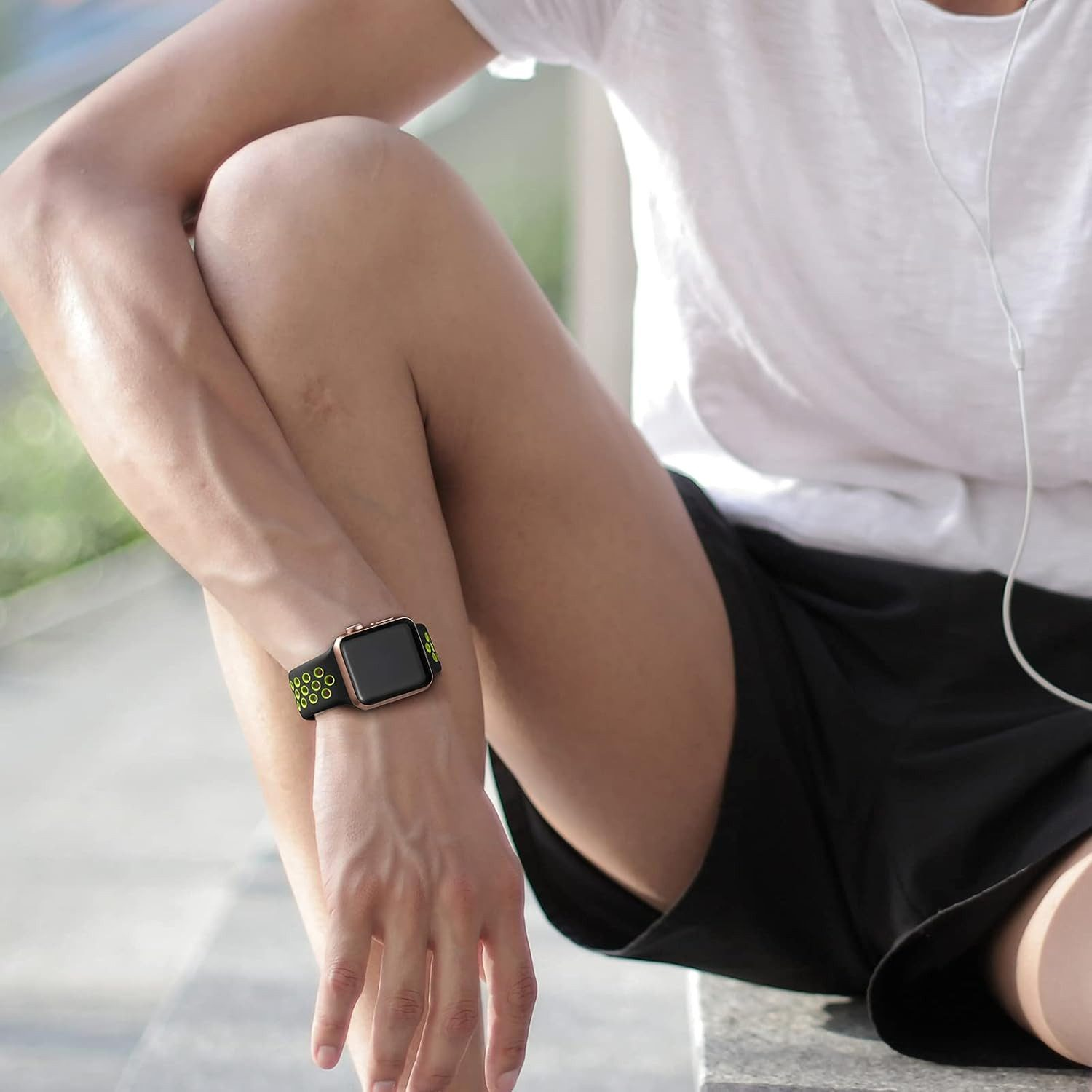 Person, die eine Smartwatch mit grünen Akzenten trägt, sitzt und trägt ein weißes Hemd und schwarze Shorts. Kopfhörerkabel sind sichtbar.