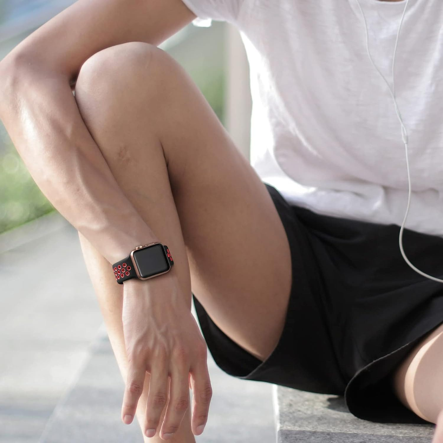 Eine Person sitzt, trägt ein weißes T-Shirt und schwarze Shorts. Sie hat eine Smartwatch mit einem schwarz-roten Band am Handgelenk.