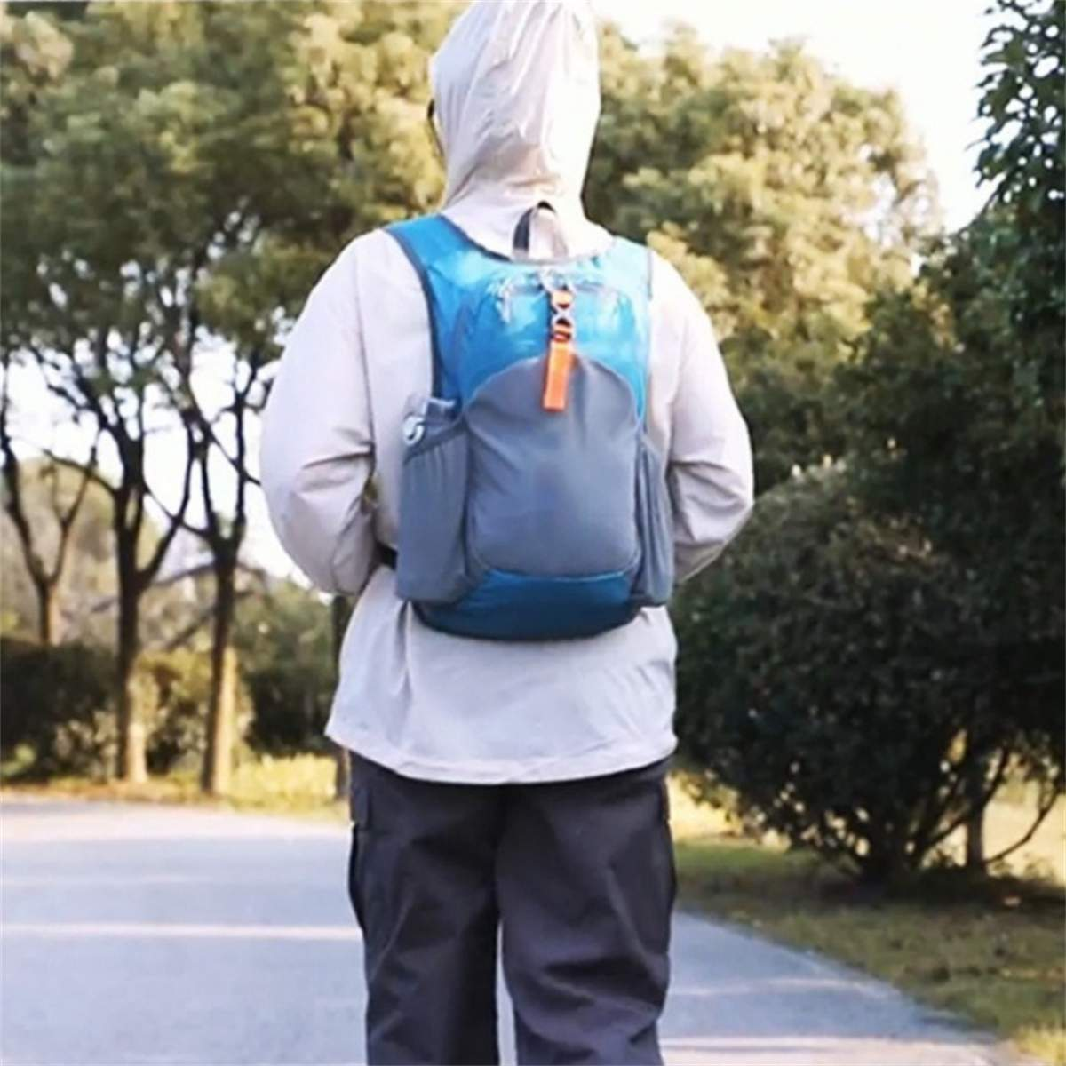 Person geht weg, trägt einen blauen Rucksack, eine Kapuzenjacke und dunkle Hosen. Bäume und Straße im Hintergrund.