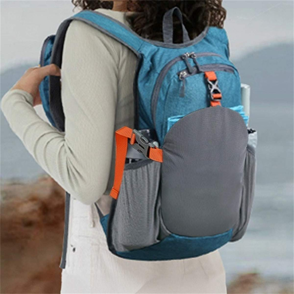 Eine Person trägt einen blauen Rucksack mit orangefarbenen Trägern. Der Rucksack hat eine graue Seitentasche. Die Person trägt ein weißes Hemd.