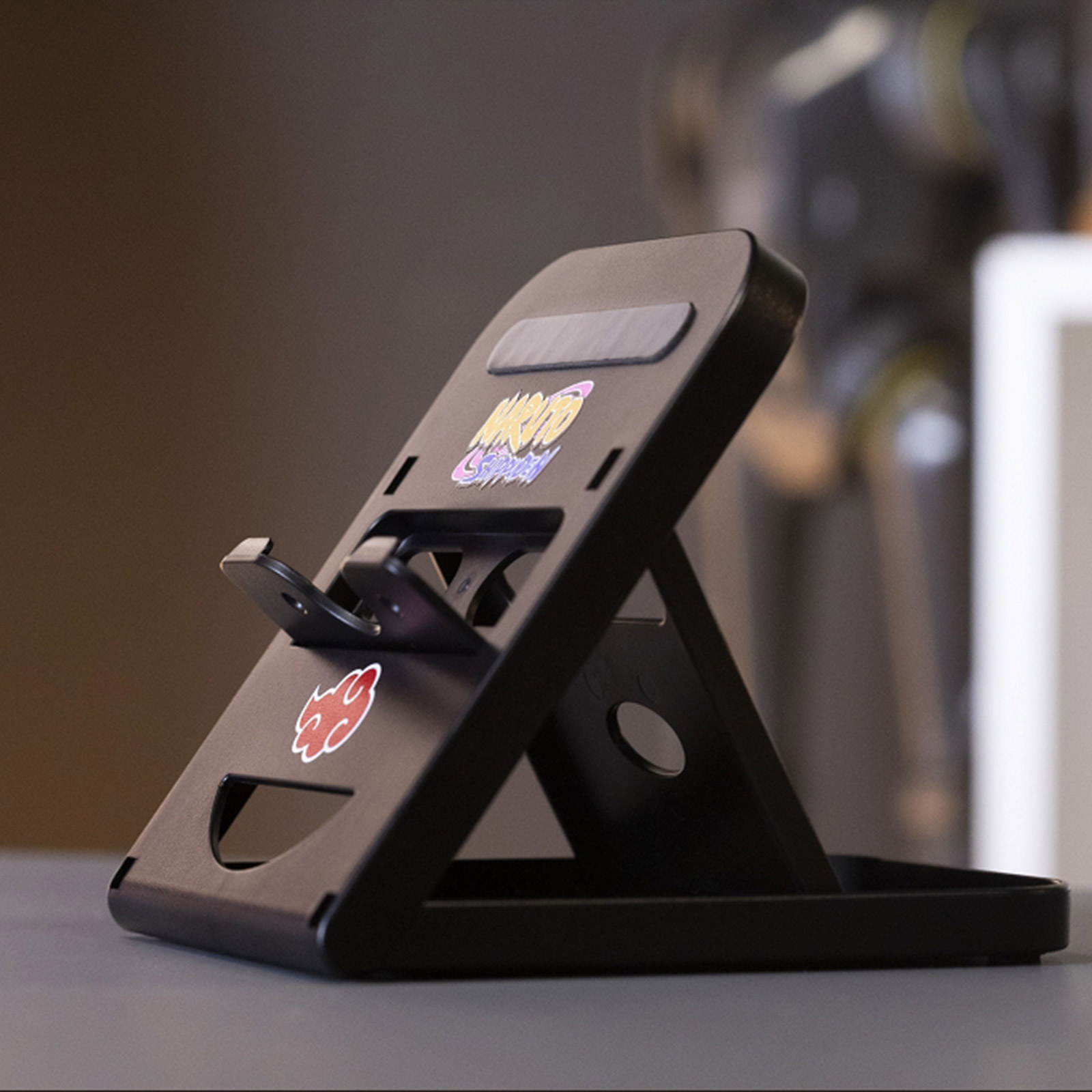 Czarny stojak na telefon z symbolami Naruto i Akatsuki. Znajduje się na szarej powierzchni z rozmytym tłem.