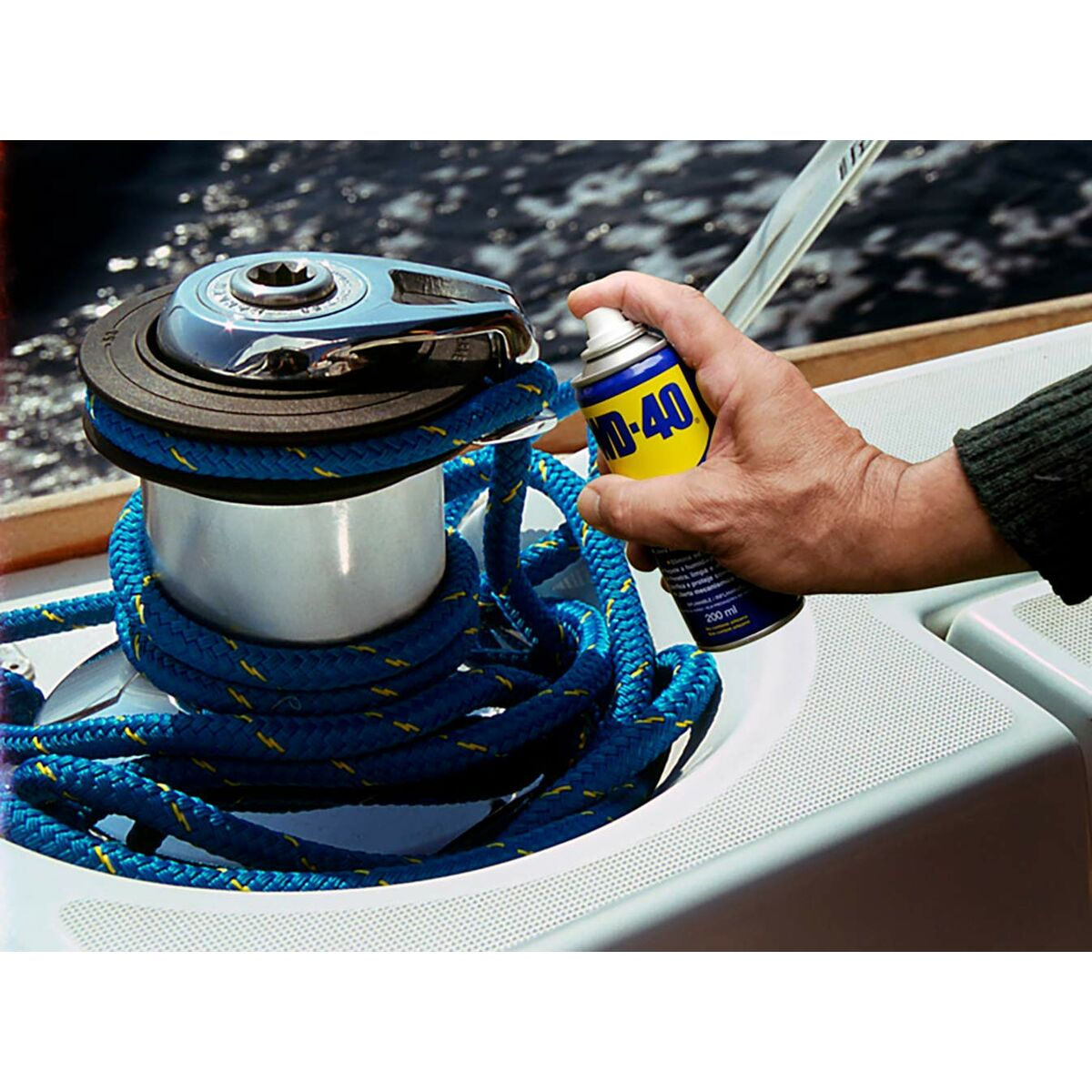 Una persona rocía WD-40 sobre un cabrestante de barco con una cuerda azul. El fondo muestra agua.