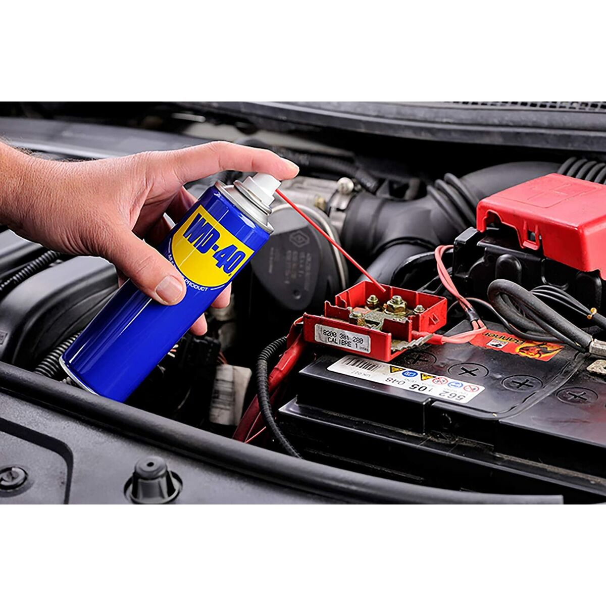 Una persona rocía WD-40 sobre una batería de coche y un motor. La lata azul se sostiene sobre el terminal rojo de la batería.