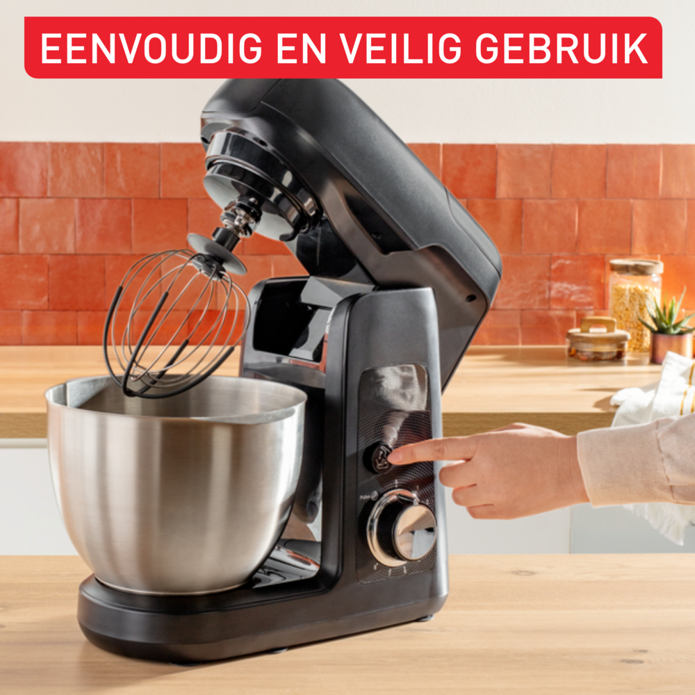 Ein schwarzer Küchenmixer mit silberner Schüssel und Schneebesen. Eine Hand drückt einen Knopf. Hintergrund: Holzoberfläche und orangefarbene Fliesen.