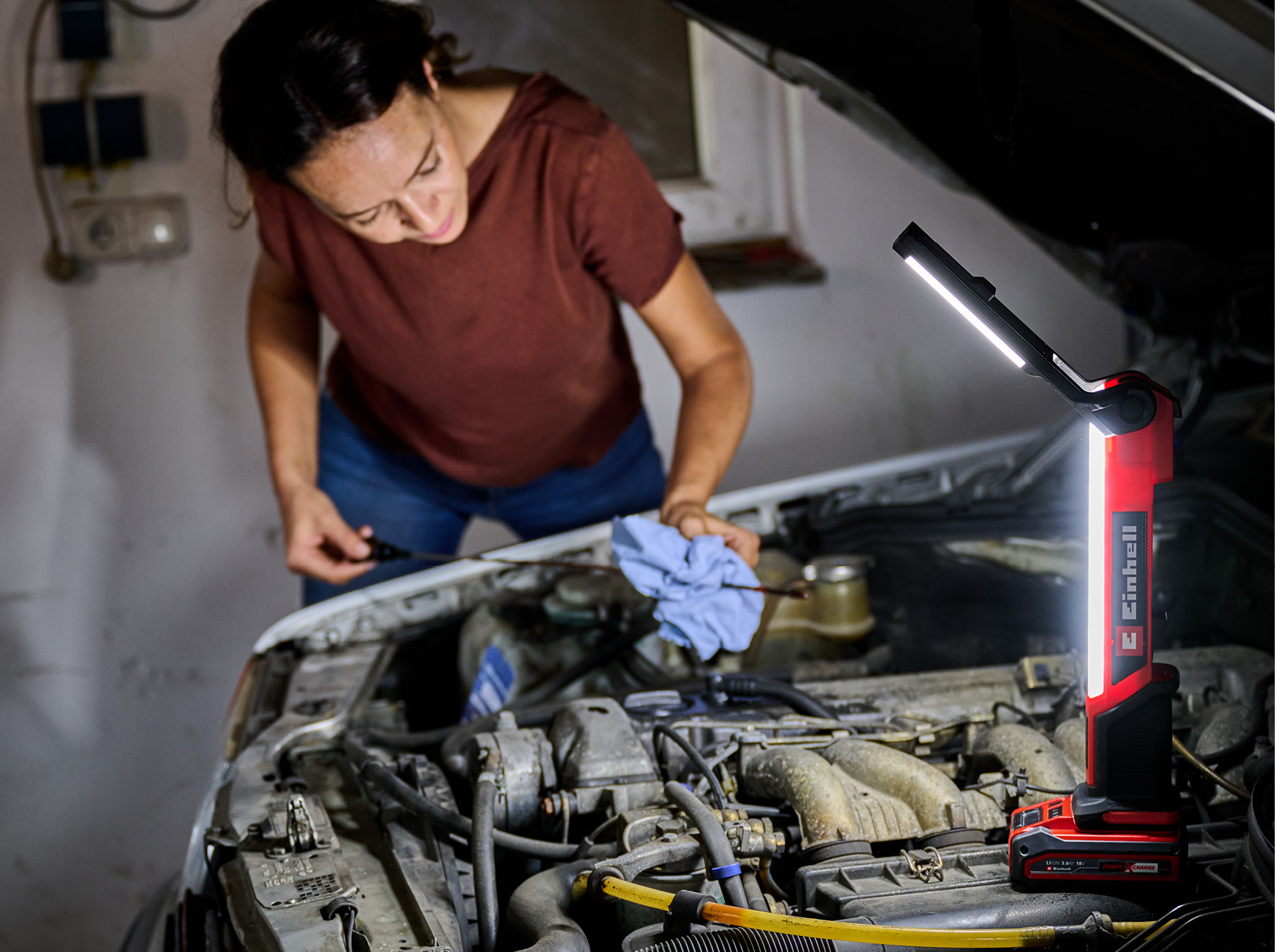 Eine Frau in einer Garage arbeitet an einem Automotor, beleuchtet von einer roten Arbeitsleuchte. Sie betrachtet einen Ölmessstab.