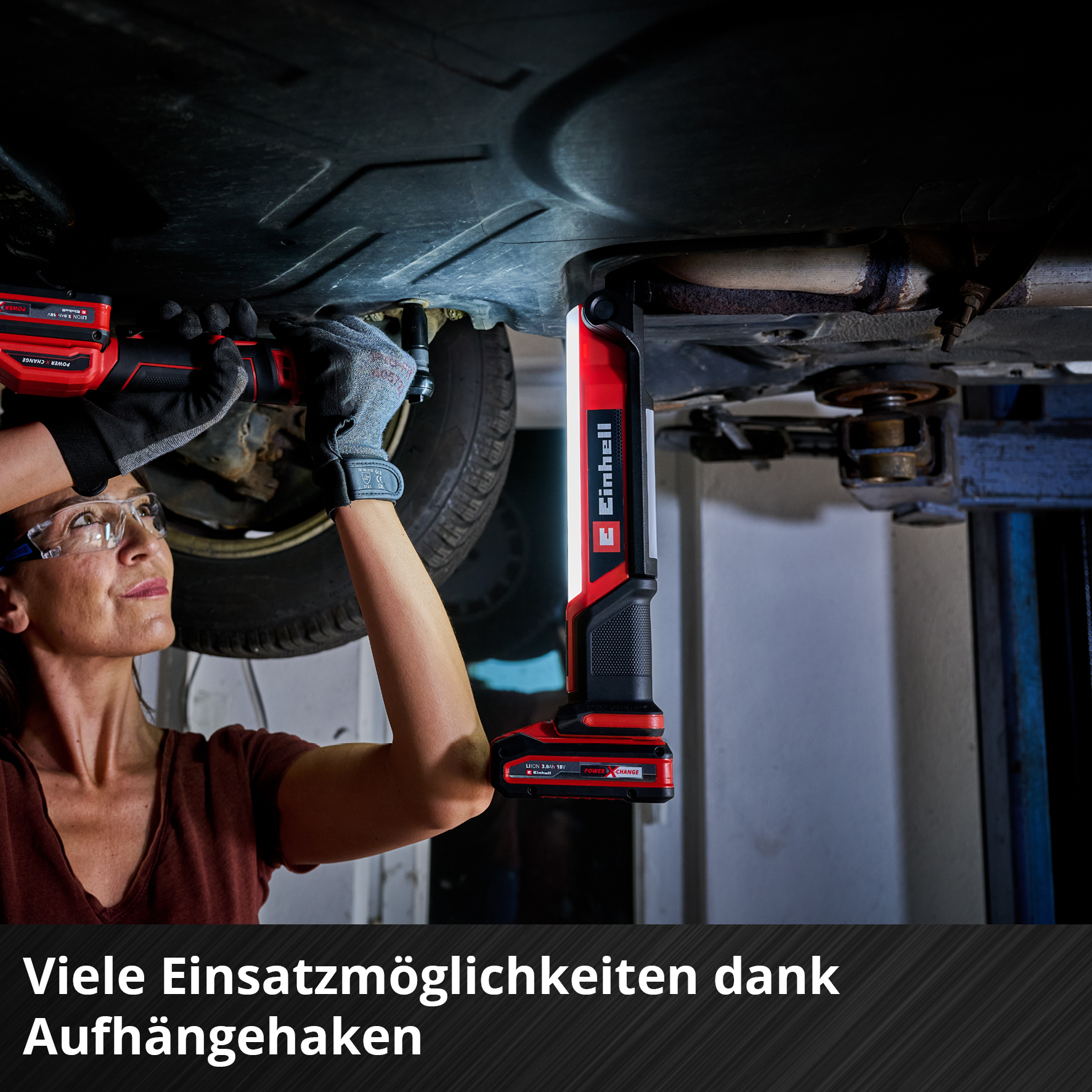 Eine Frau in einer Werkstatt benutzt ein rot-schwarzes Arbeitslicht unter einem Auto. Schutzbrille und Handschuhe sind ebenfalls zu sehen.