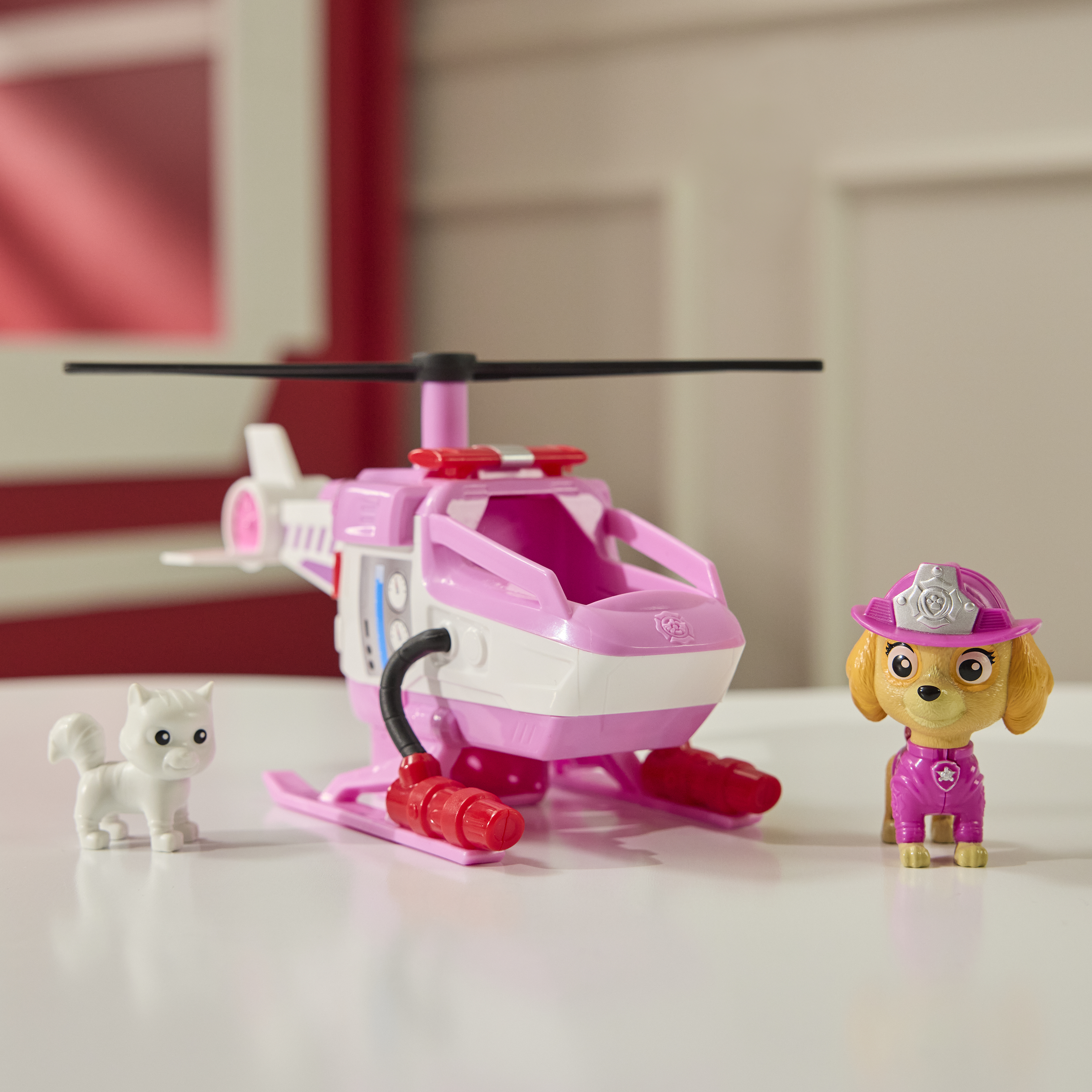 Ein rosa Helikopter-Spielzeug mit einer weißen Katze und einer Skye-Figur auf einer weißen Oberfläche. Hintergrund zeigt eine rote Wand und eine weiße Tür.