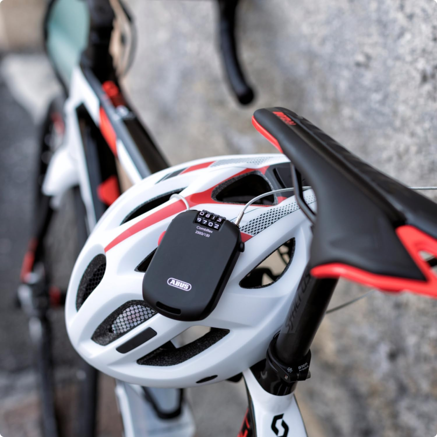 Biały kask rowerowy z czerwonymi paskami i czarną blokadą szyfrową. Widoczne czarne siodełko i kierownica.