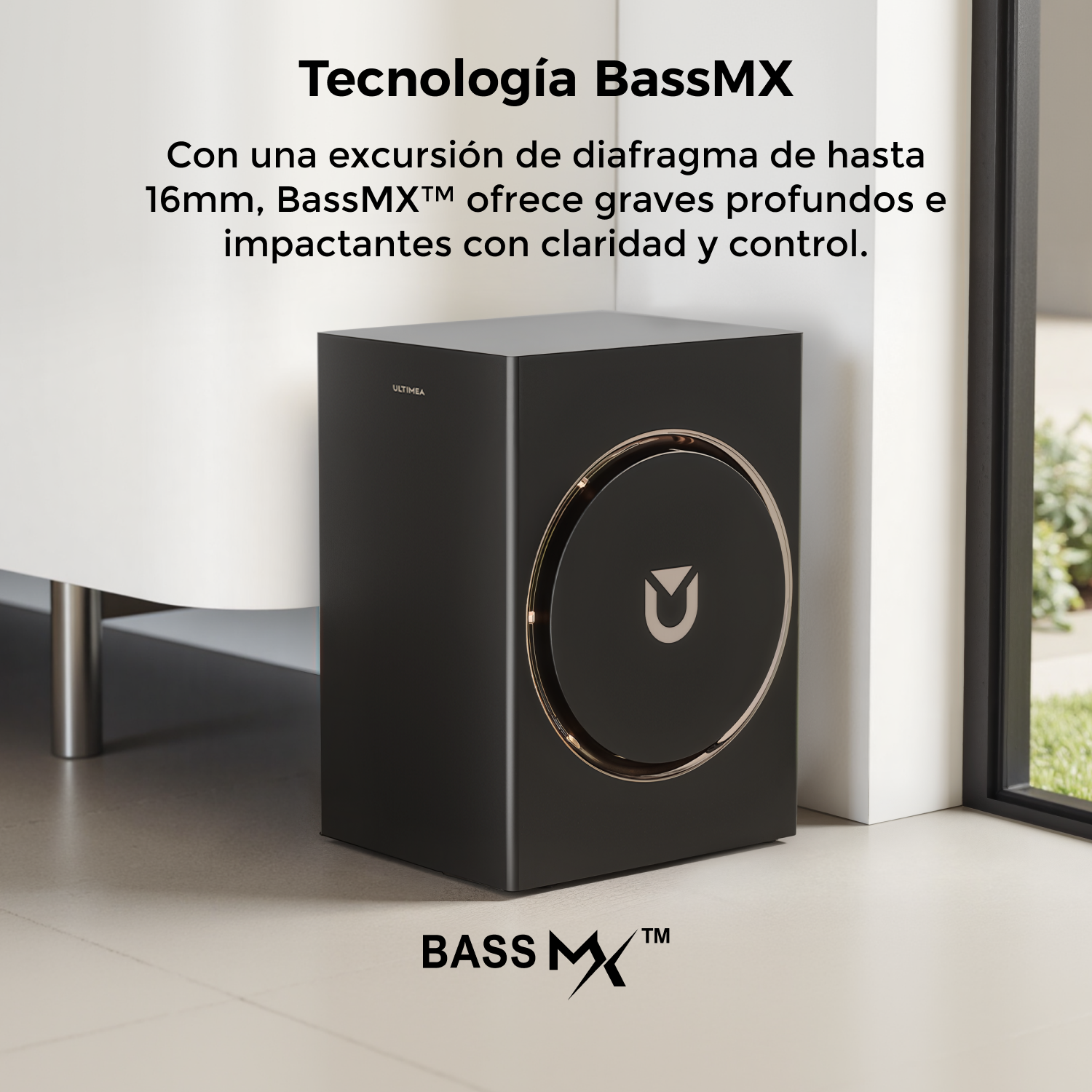 Un altavoz BassMX negro con un anillo y logo dorados, colocado cerca de una pared y ventana blancas, texto en español.