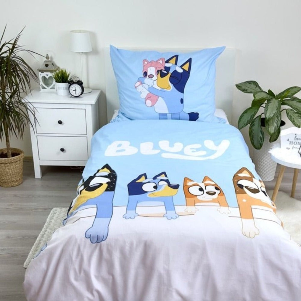 Bettwäsche mit Bluey-Figuren. Das Set umfasst einen Kissenbezug und eine Bettdecke mit Bluey-Designs. Ein Nachttisch mit Uhr steht neben dem Bett.