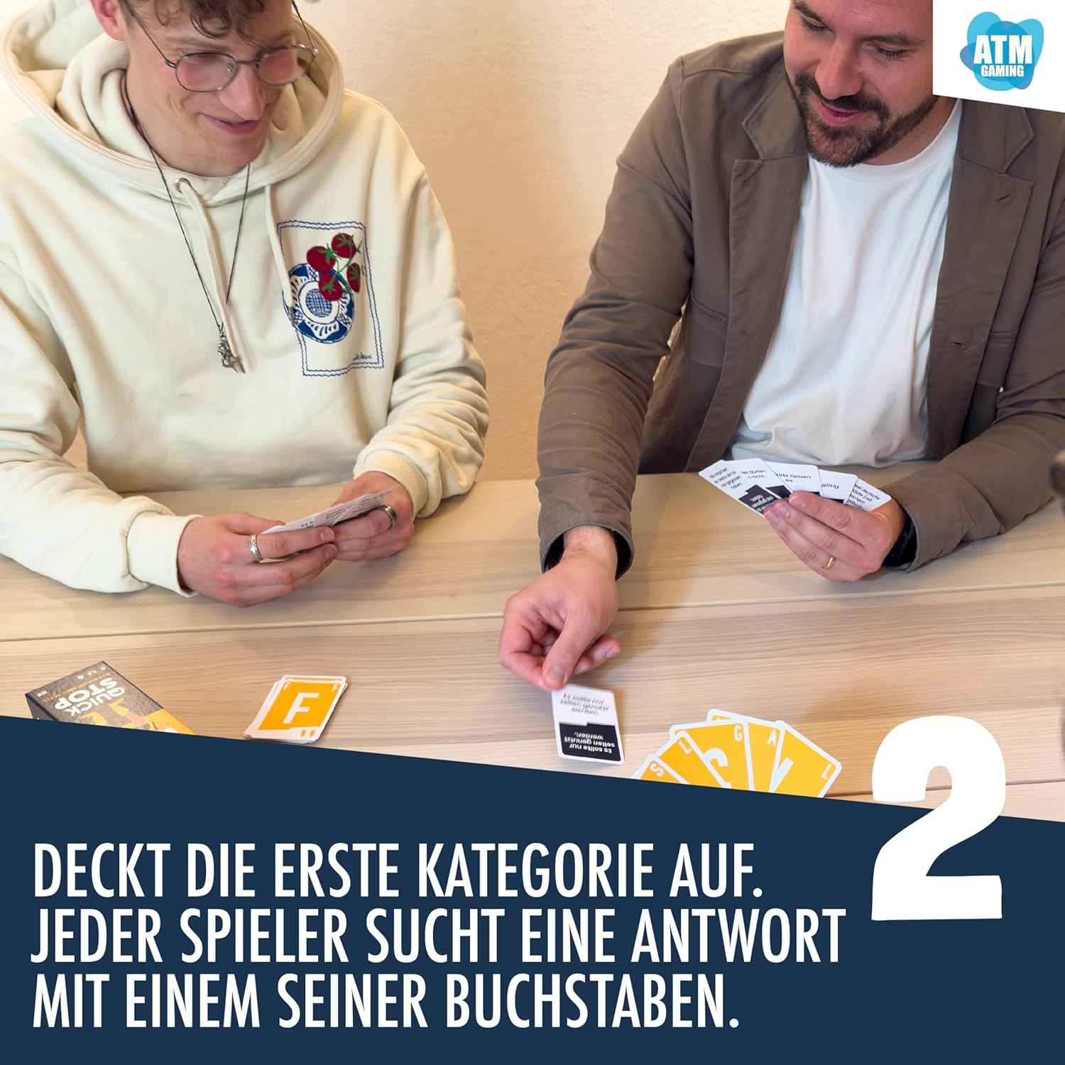 Deux hommes jouant à un jeu de cartes, texte en allemand. Cartes avec lettres et cartes de jeu sur la table. Logo "ATM Gaming" visible.