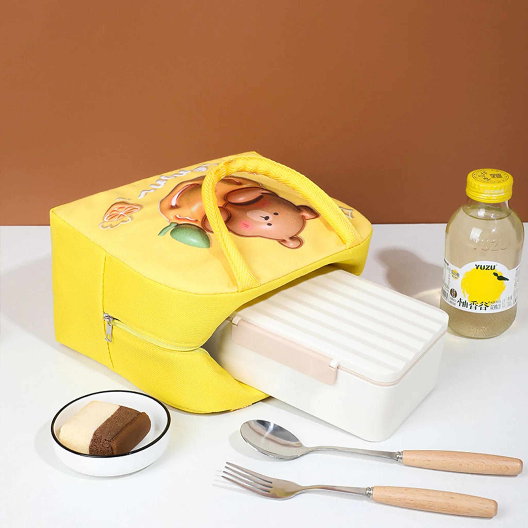 Gelbe Lunchtasche mit Bär, Brotdose, Getränkeflasche, Kuchen, Löffel und Gabel. Brauner Hintergrund.