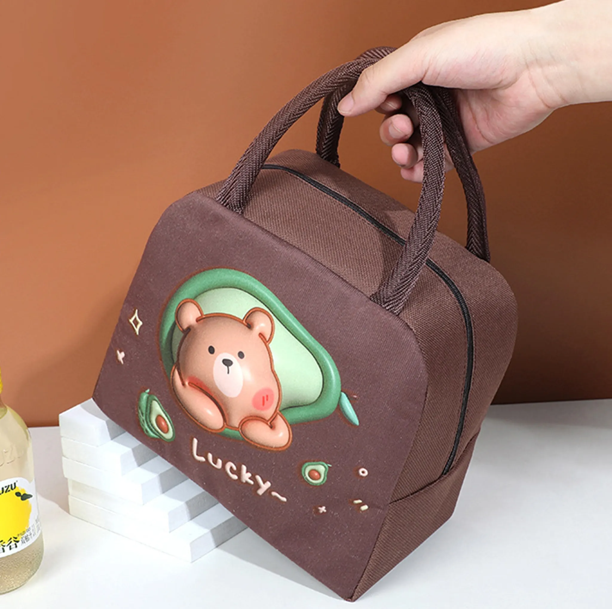 Eine braune Lunch-Tasche mit einem Bären-Avocado-Design wird von einer Hand gehalten. Der Hintergrund ist braun und weiß.
