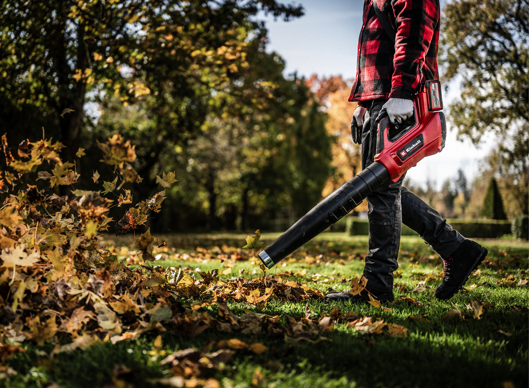 Une personne en chemise à carreaux rouge utilise un souffleur de feuilles pour nettoyer les feuilles mortes sur une pelouse d'automne.