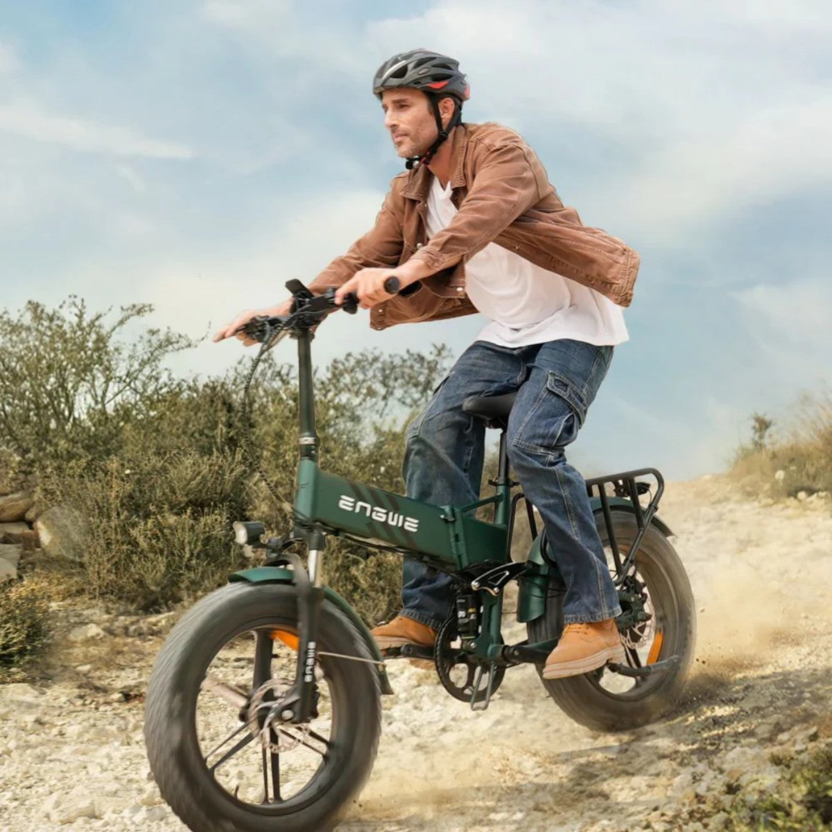 Un uomo guida una bici elettrica verde su una strada sterrata. Indossa un casco, una giacca marrone e jeans. Terreno polveroso e cielo sullo sfondo.