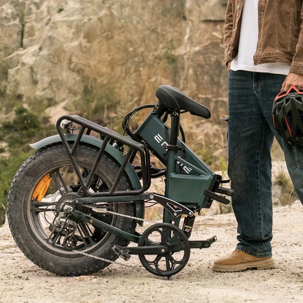 Una e-bike accanto a un uomo in jeans e giacca marrone che tiene un casco. Uno sfondo roccioso dietro di loro.