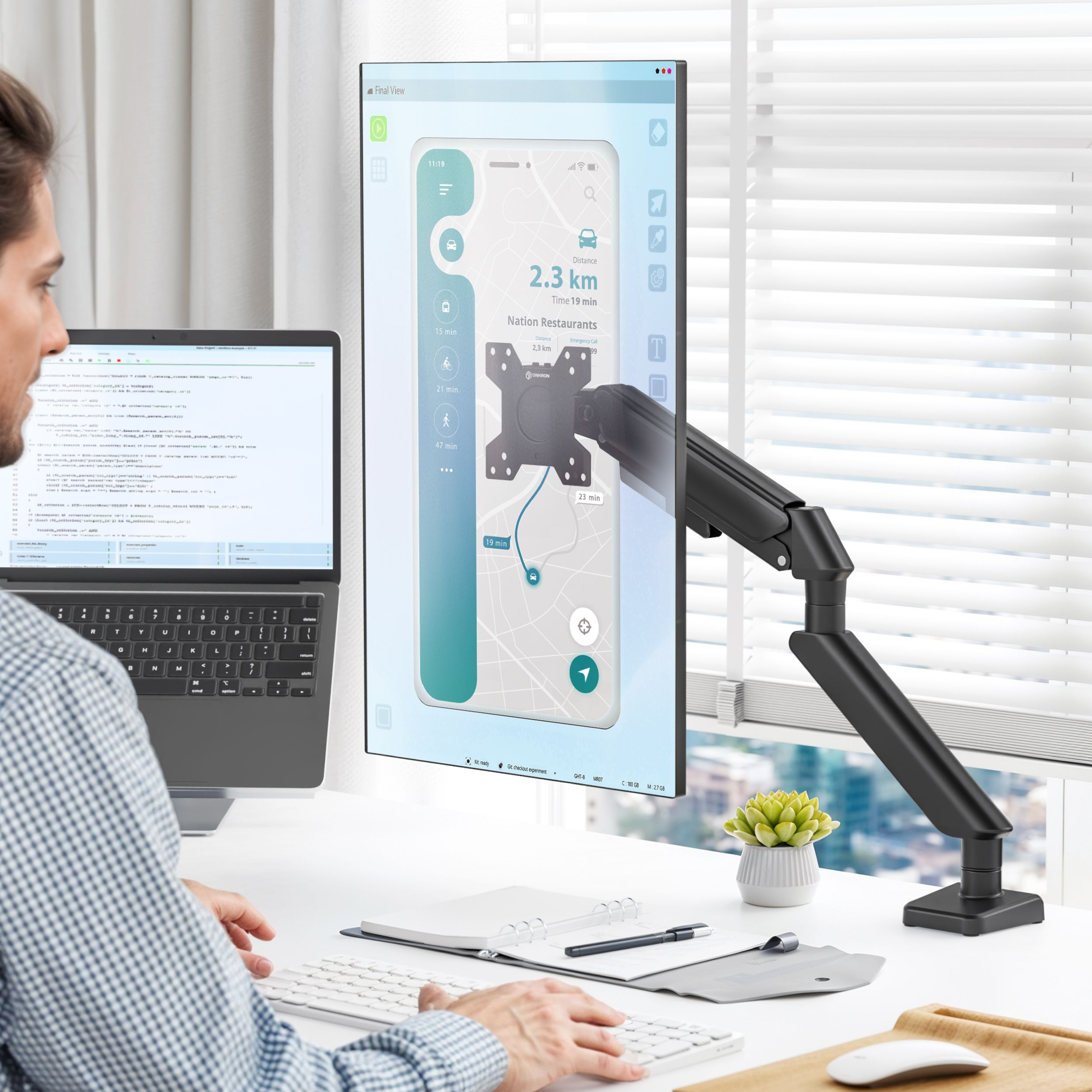 Un hombre trabaja en un portátil y un monitor que muestra un mapa. Teclado, ratón y bloc de notas en el escritorio.