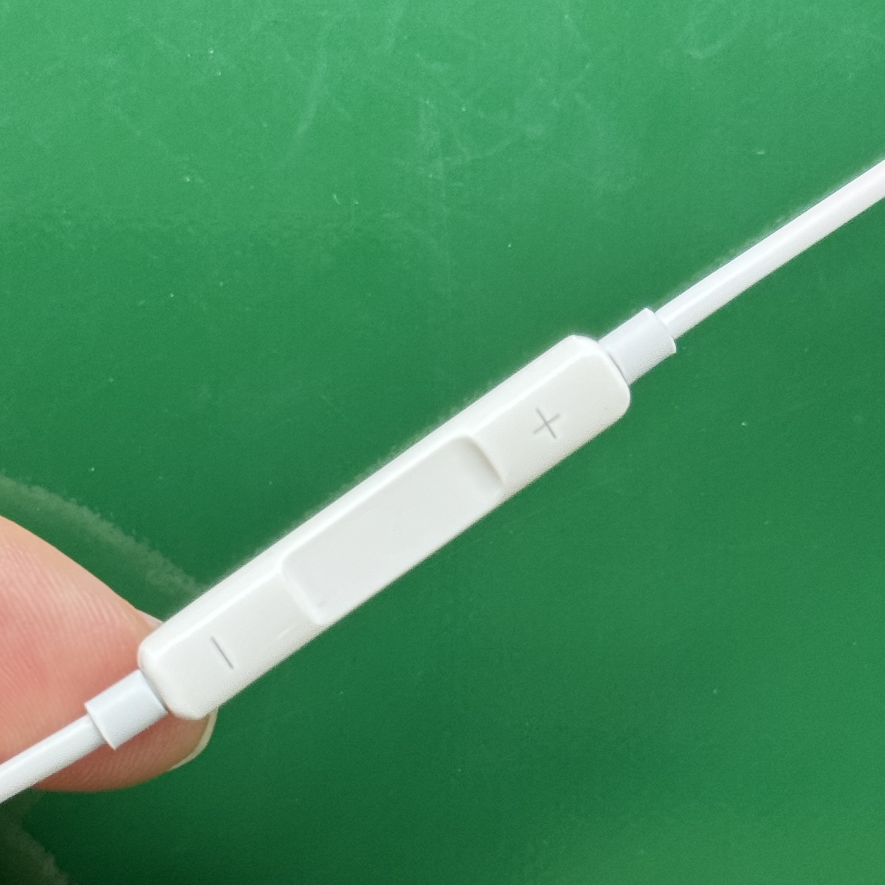 Weißes Kopfhörer-Bedienelement mit Plus- und Minuszeichen auf grünem Hintergrund. Ein Finger hält die Steuerung.