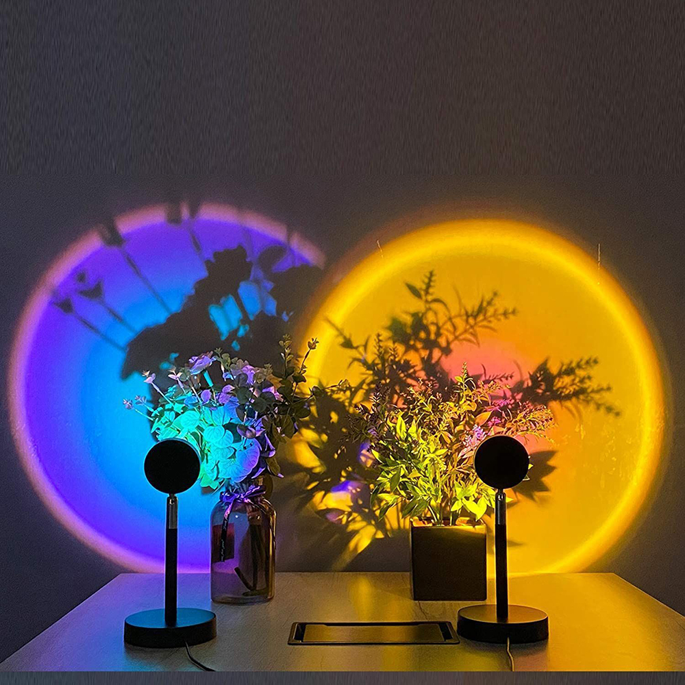 Zwei Sonnenuntergangslampen projizieren Lichtkreise auf eine Wand. Pflanzen und Lampen stehen auf einem Tisch. Eine Lampe hat einen blauen Kreis, die andere gelb.