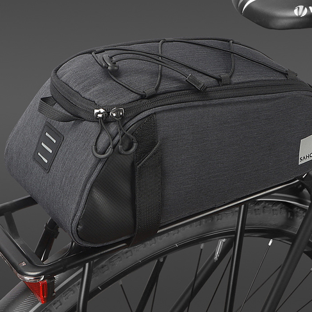 Eine schwarze Fahrradtasche ist auf einem Gepäckträger montiert. Die Tasche hat einen Reißverschluss, einen Reflektorstreifen und elastische Kordeln. Der Hintergrund ist dunkel.