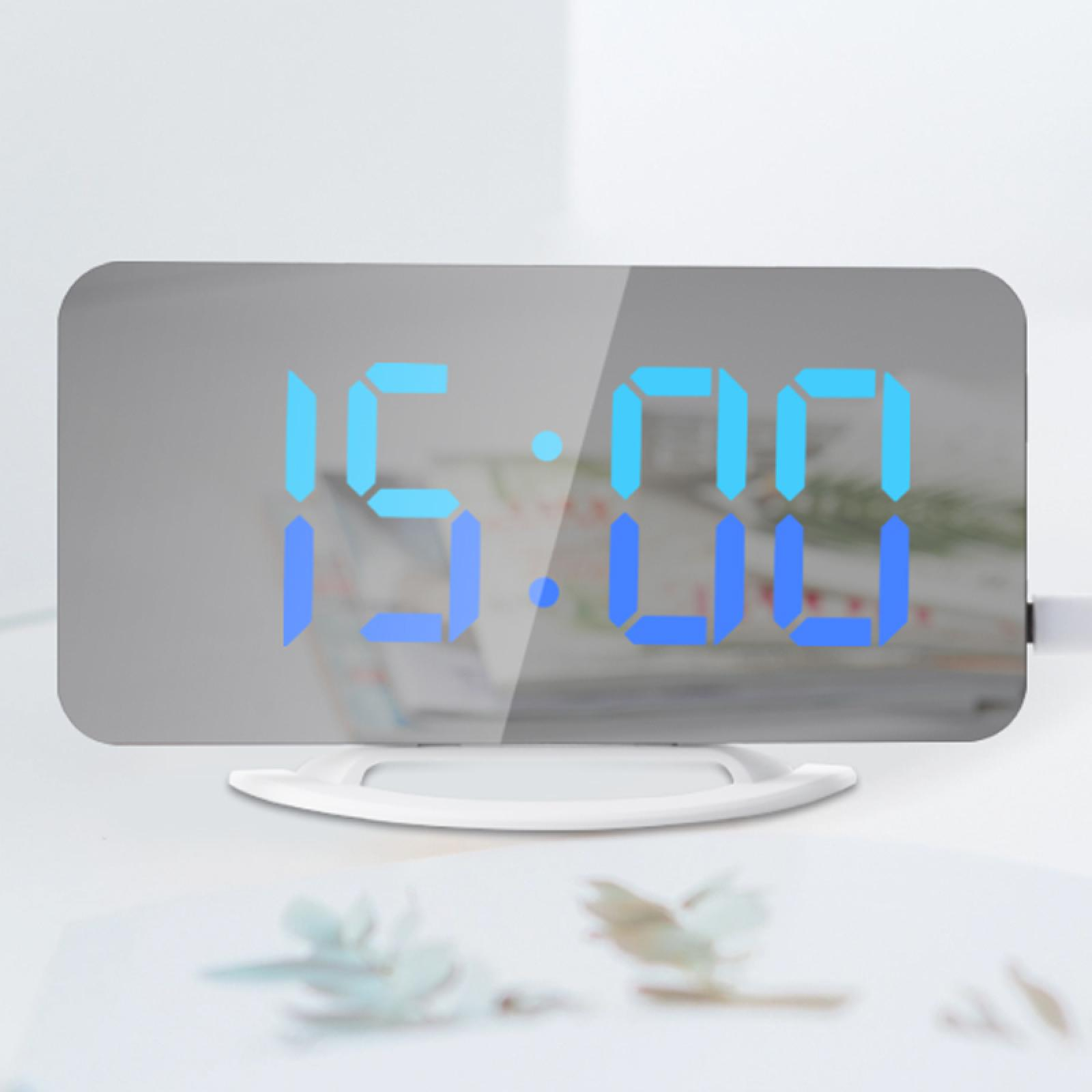 Eine Digitaluhr zeigt 15:00 in Blau auf einer reflektierenden Oberfläche an. Sie steht auf einem weißen Ständer, mit verschwommenen Blumen im Vordergrund.