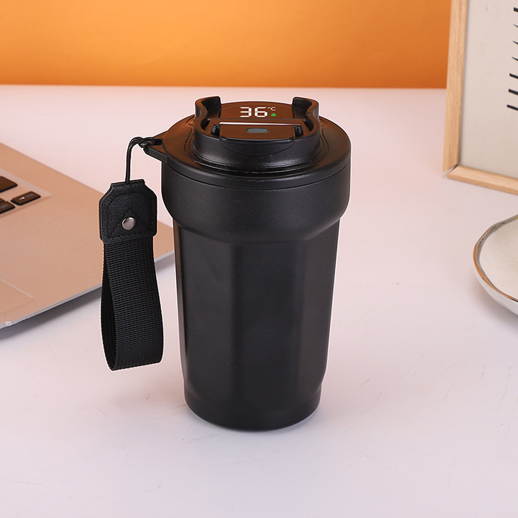 Schwarzer intelligenter Thermobecher mit digitaler Temperaturanzeige. Ein schwarzer Riemen hängt zur Seite. Laptop im Hintergrund.