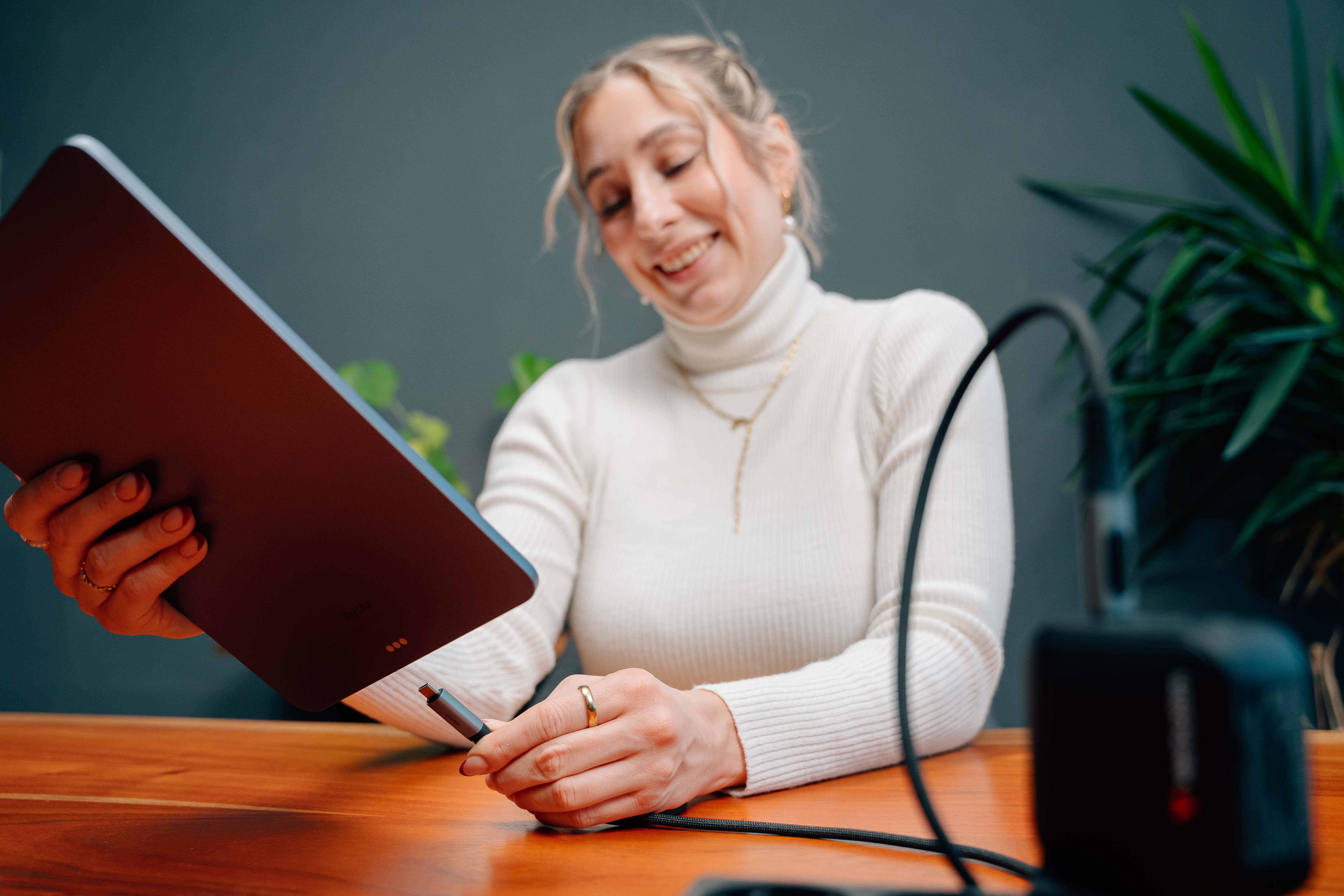 Femme tenant une tablette, souriante. Elle porte un pull blanc et des bijoux en or. Un câble est branché sur la tablette.