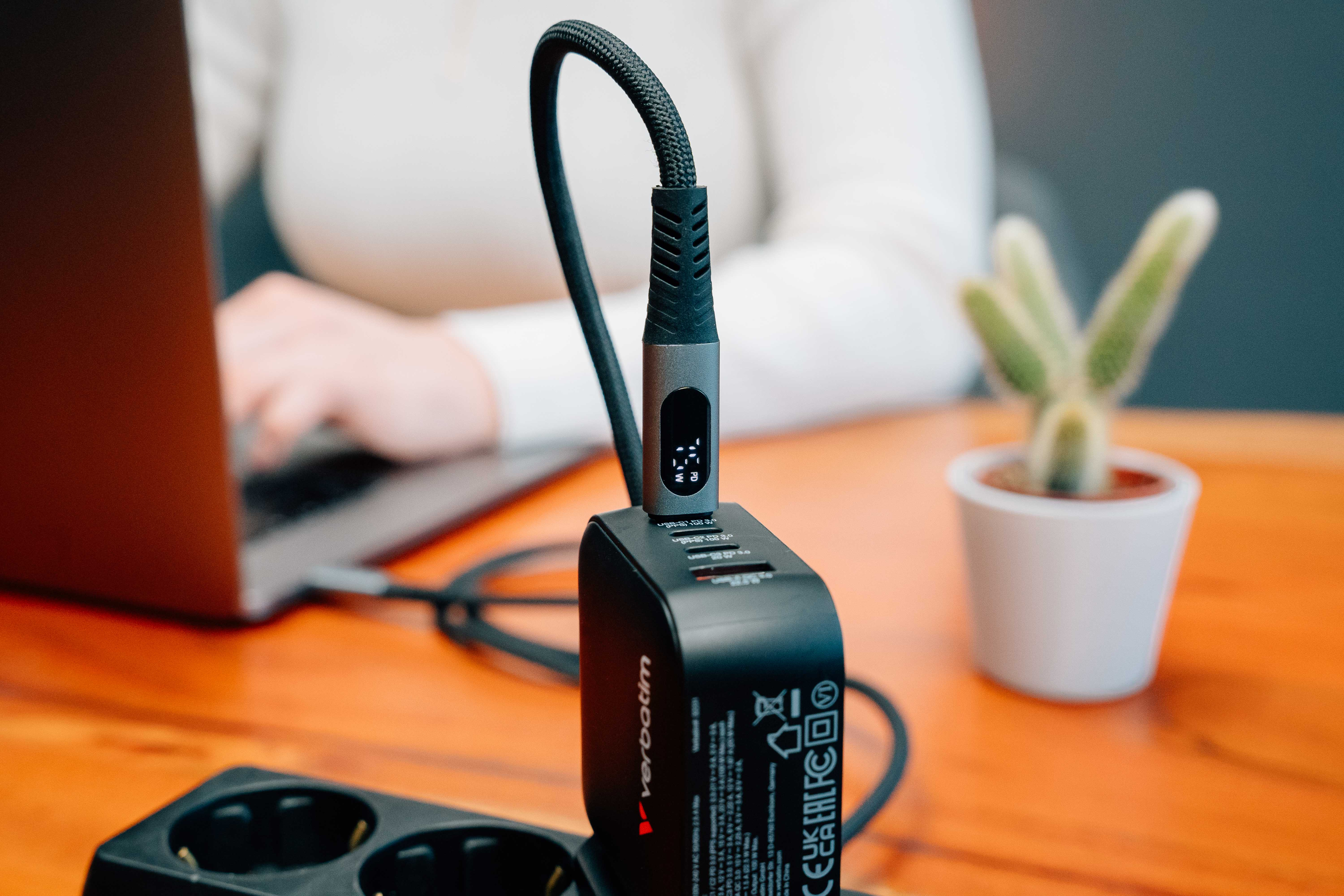 Un chargeur Verbatim noir avec câble connecté, un ordinateur portable et un petit cactus en pot blanc sur une table orange.