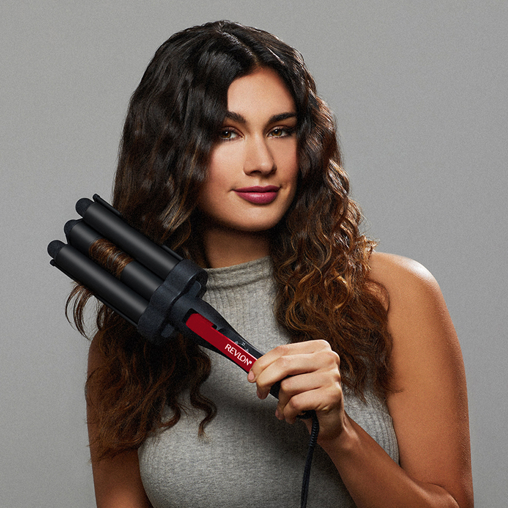 Eine Frau hält einen Revlon-Haarwellenstab. Sie hat welliges braunes Haar. Der Hintergrund ist einfarbig grau.