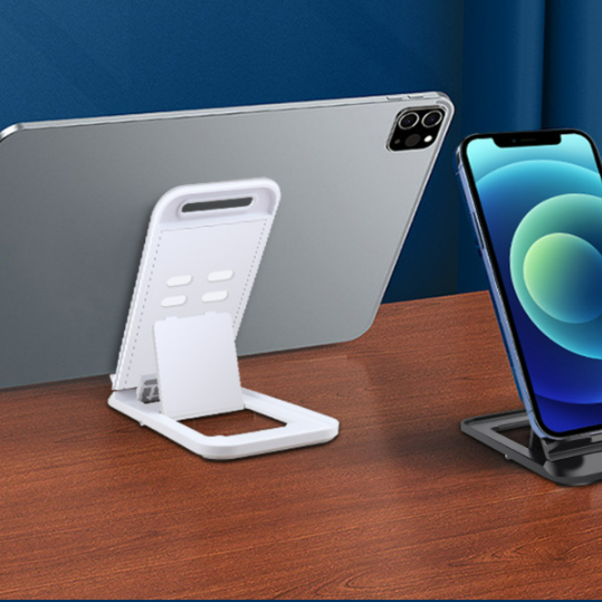 Tablet i smartfon na stojakach. Tablet jest szary, na białym stojaku. Smartfon jest niebieski, na ciemnym stojaku. Drewniany stół i niebieskie tło.
