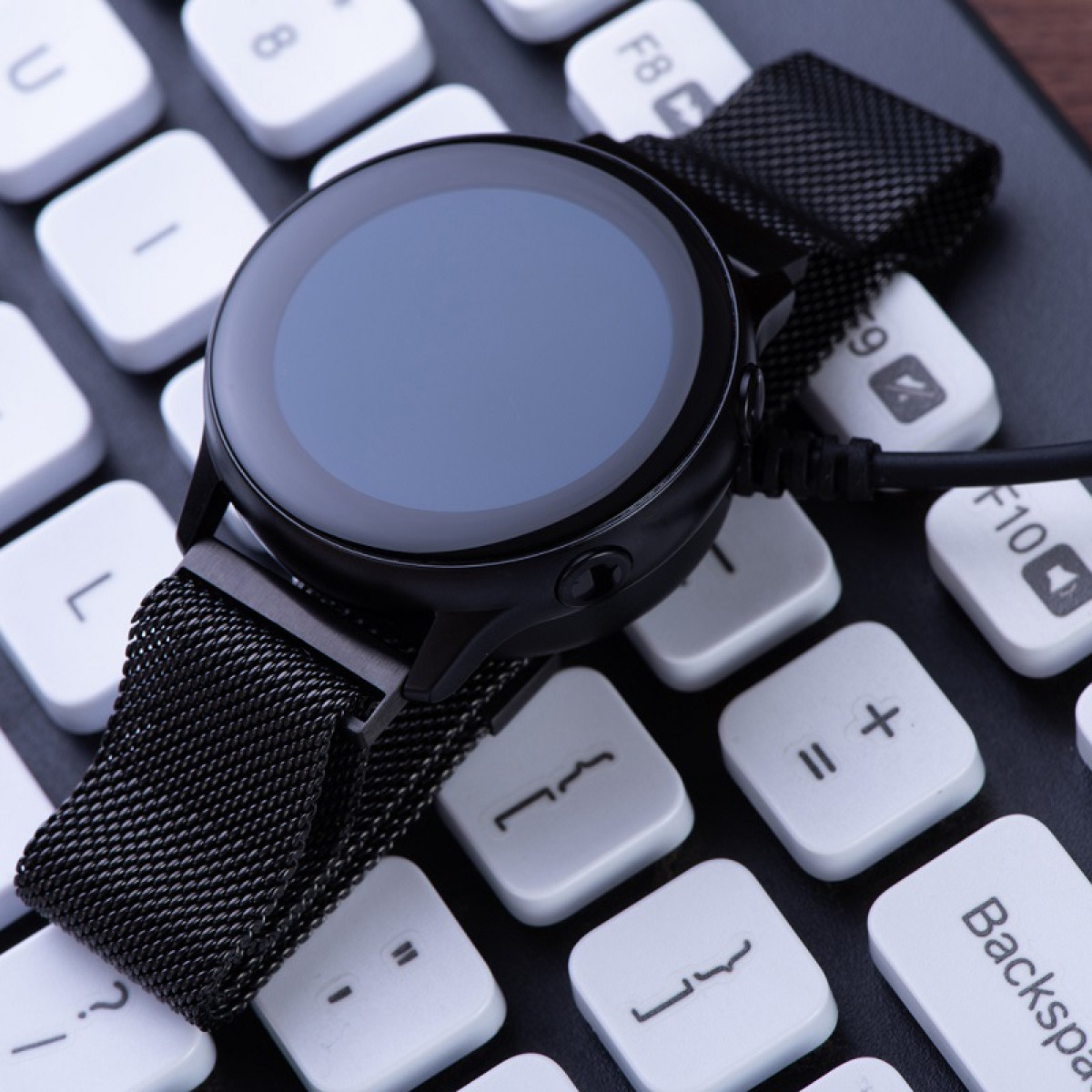 Czarny smartwatch na klawiaturze. Zegarek jest okrągły, z ciemnym ekranem i siatkowym paskiem. Klawisze klawiatury są białe.