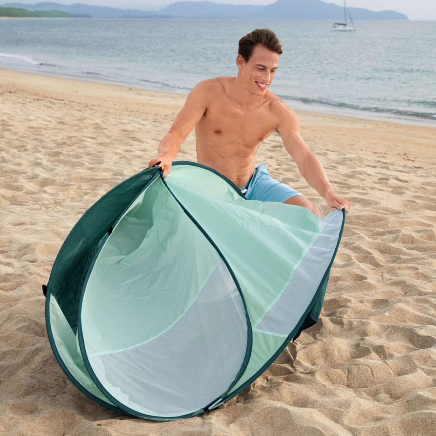 Een man op het strand zet een zeegroene tent op. Hij draagt een blauwe korte broek. De oceaan en een zeilboot zijn op de achtergrond.