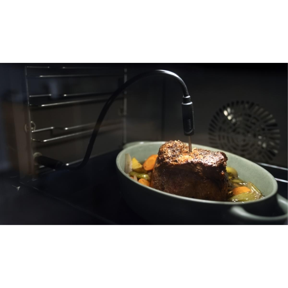 Een braadstuk in een oven met een temperatuursonde. Groenten omringen het vlees in een ovale schaal. De ovendeur is open.