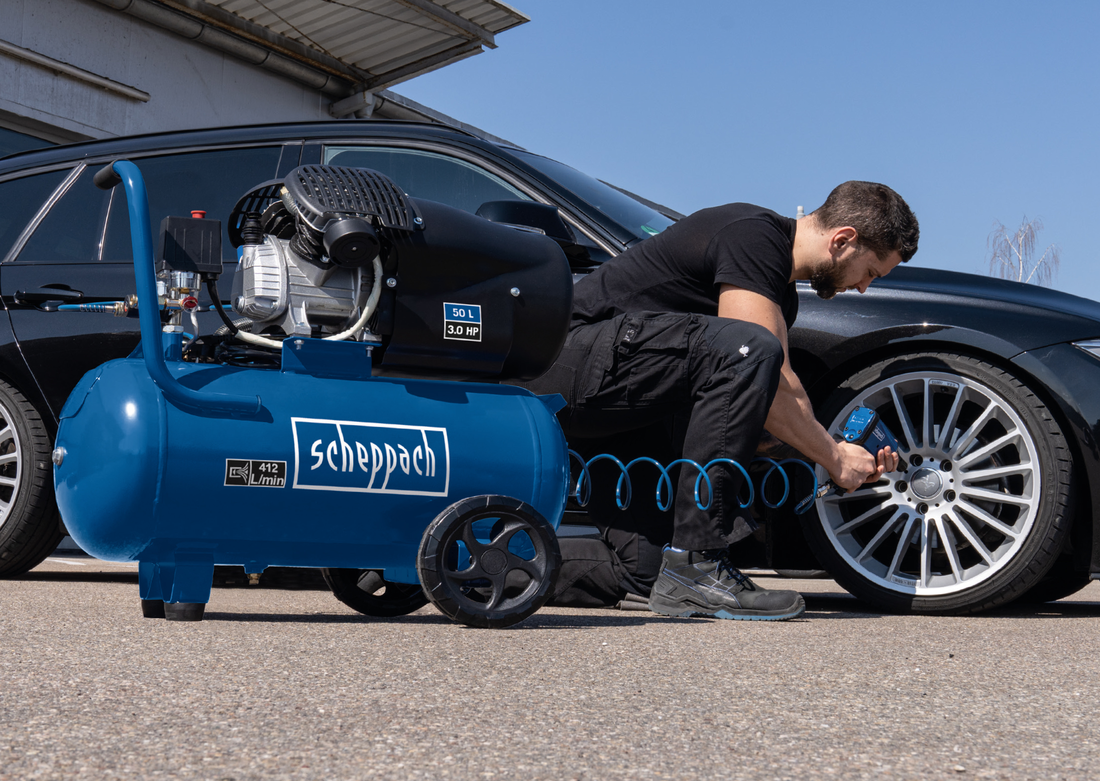 Un uomo gonfia un pneumatico di un'auto con un compressore blu. L'auto è nera e il cielo è sereno.