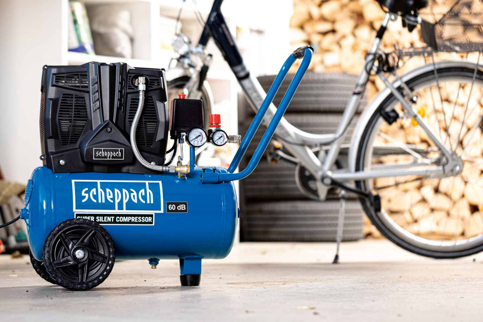 Un compressore d'aria Scheppach blu con componenti e ruote nere, una bicicletta e legna da ardere sullo sfondo.