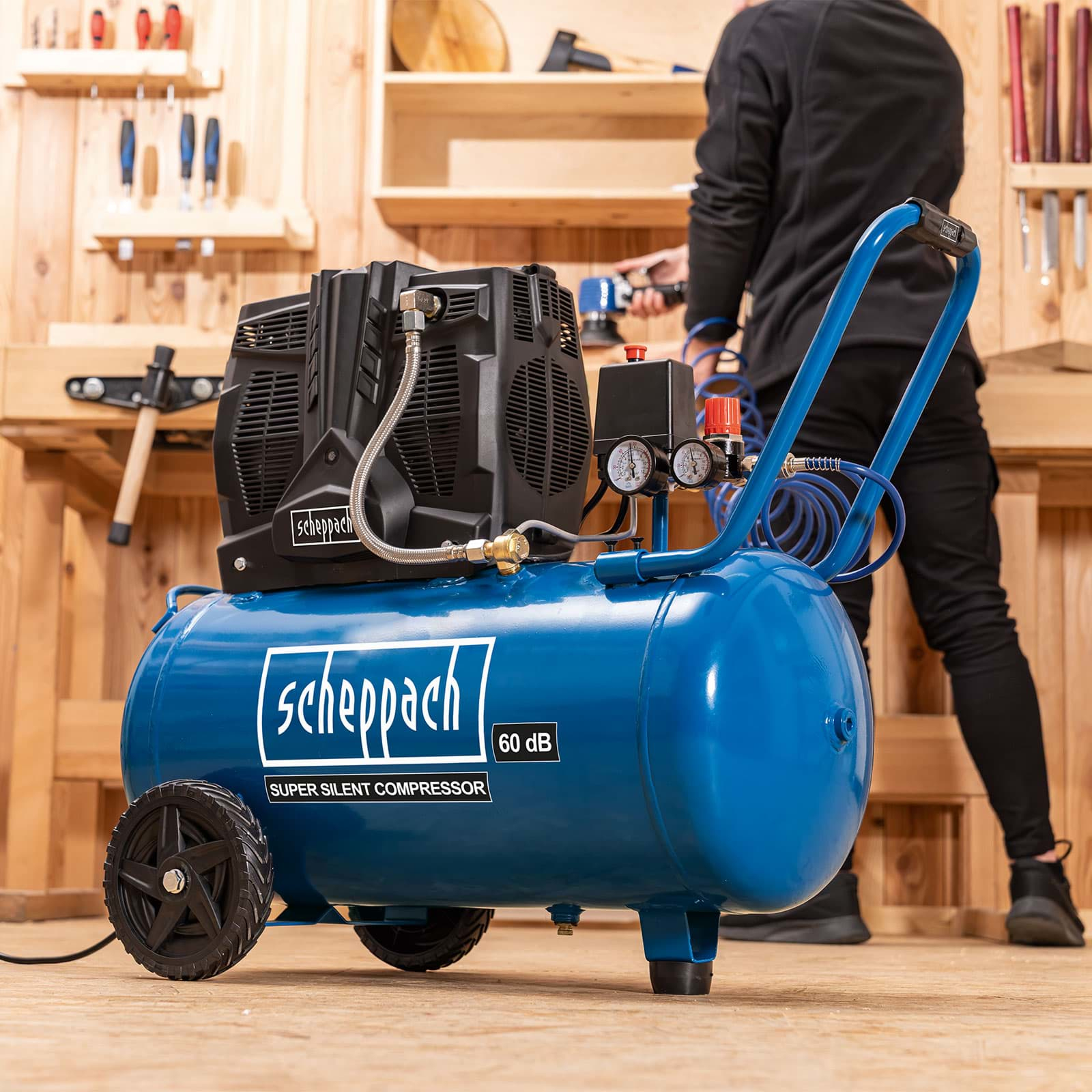 Un compressore d'aria Scheppach blu con componenti neri, ruote e una persona che lavora sullo sfondo in un'officina in legno.