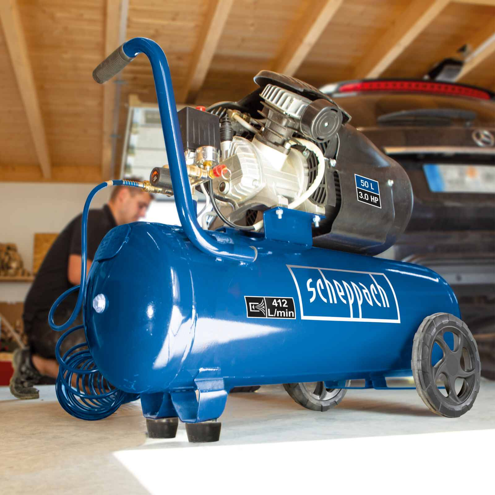 Un compressore d'aria blu con ruote, maniglia e motore nero. Una persona sullo sfondo.