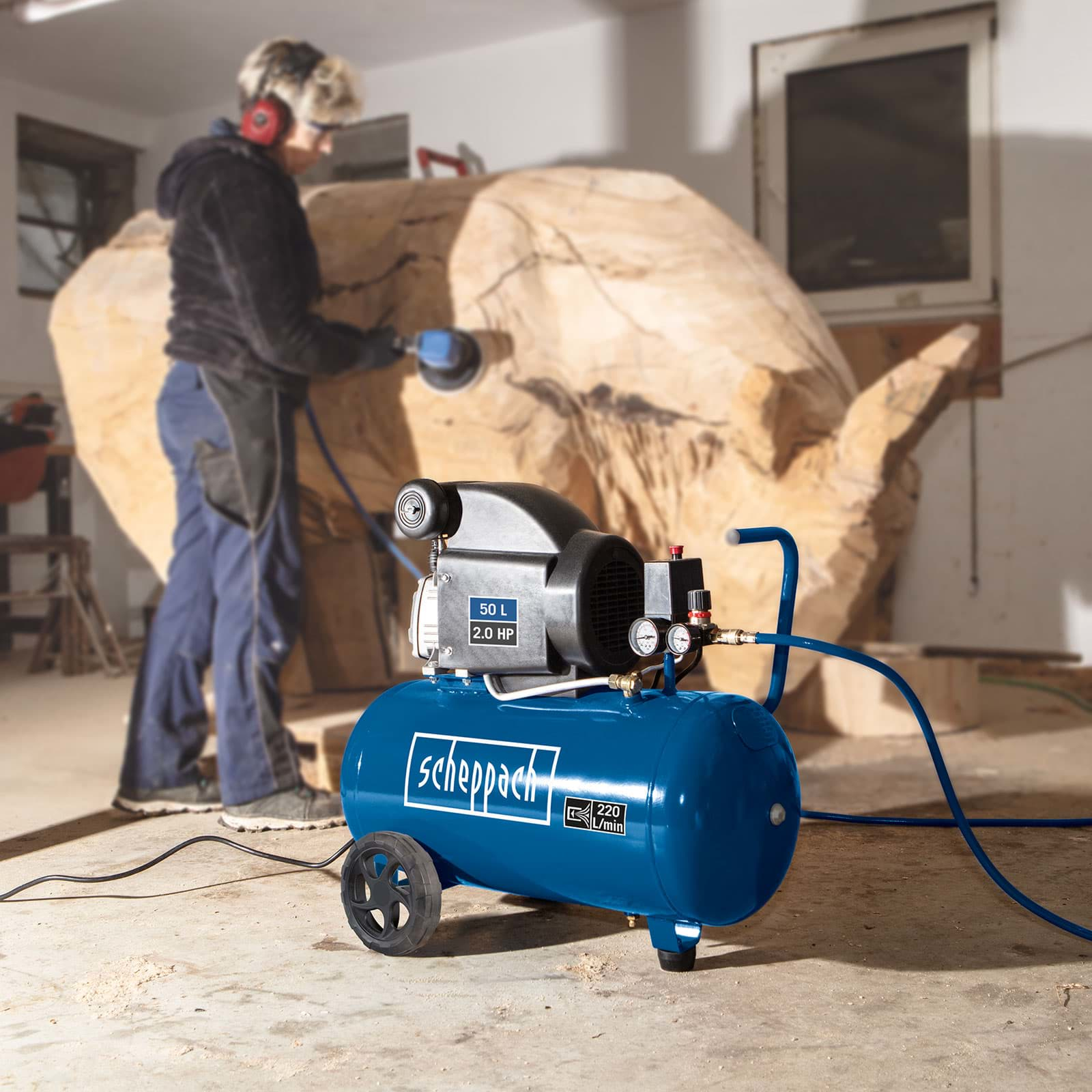 Una persona scolpisce legno con una levigatrice. Un compressore d'aria blu è in primo piano. La scena si svolge in un'officina.