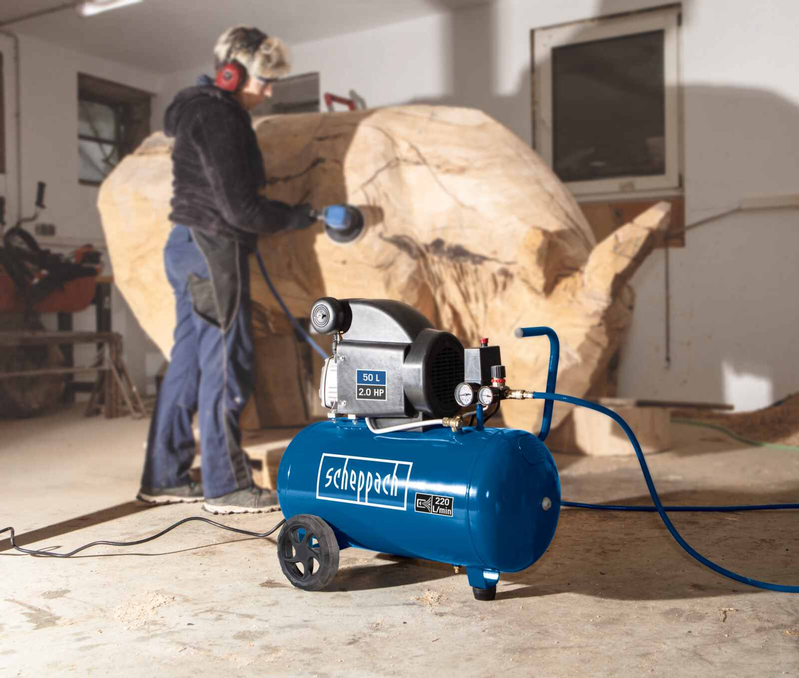 Una persona intaglia il legno con una levigatrice. Un compressore d'aria blu con ruote è sul pavimento. L'officina ha una finestra.