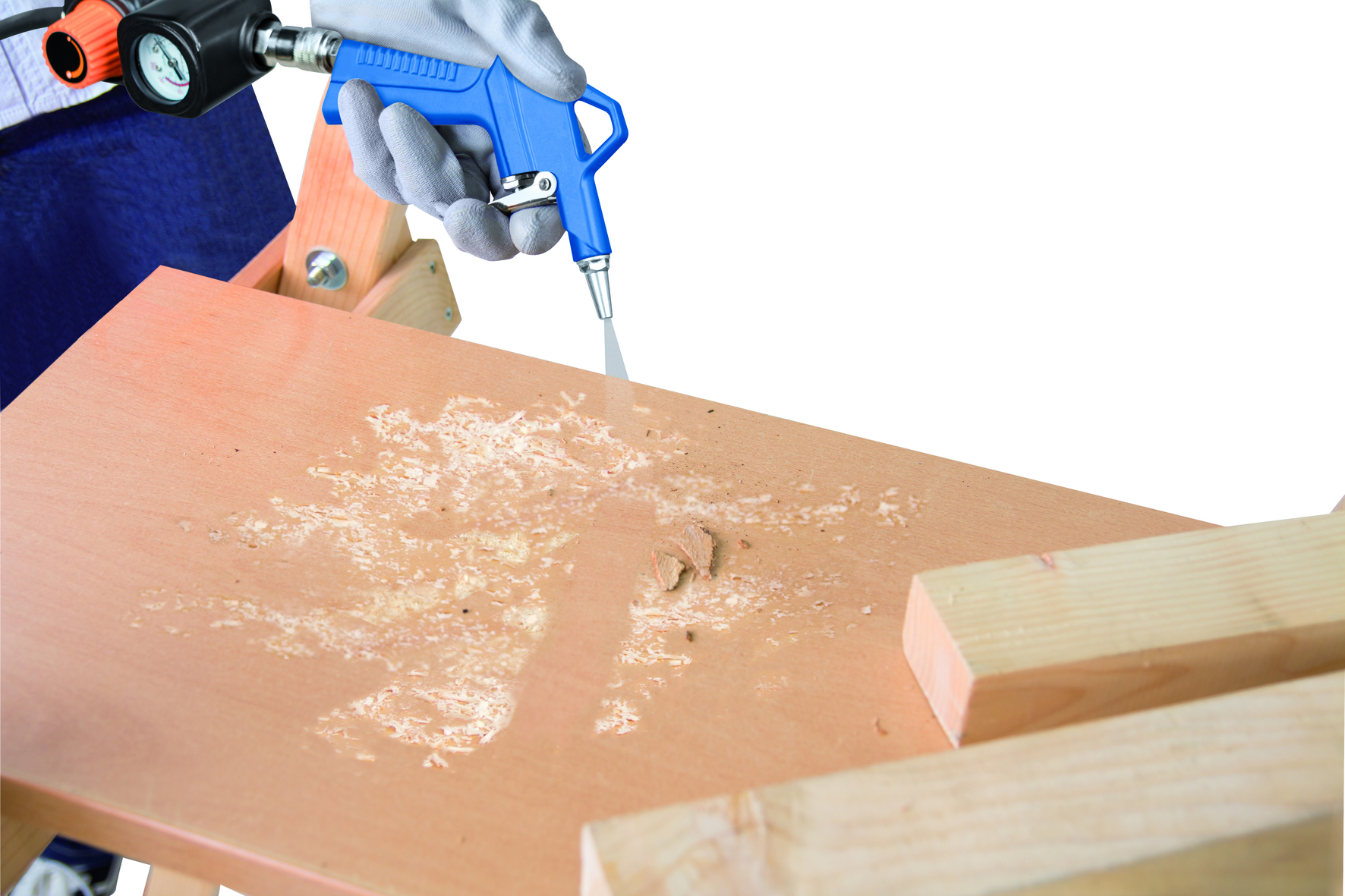 Una mano guantata tiene una pistola ad aria blu, soffiando via i detriti da una superficie di legno. Sono visibili anche un manometro e dei supporti in legno.