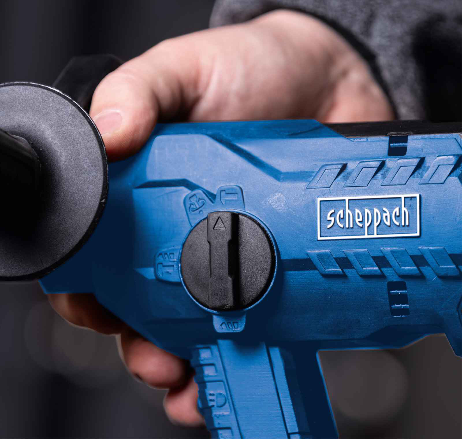 Eine Hand hält einen blauen Scheppach-Bohrer. Der Bohrer hat einen schwarzen Griff und ein schwarzes Zifferblatt. Der Hintergrund ist unscharf.