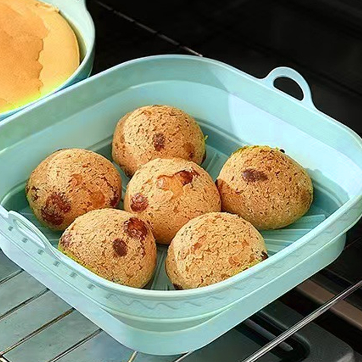 Sechs goldbraune Brötchen in einer hellblauen quadratischen Backform. Ein weiterer Behälter mit einem teilweise gebackenen Brötchen ist sichtbar.