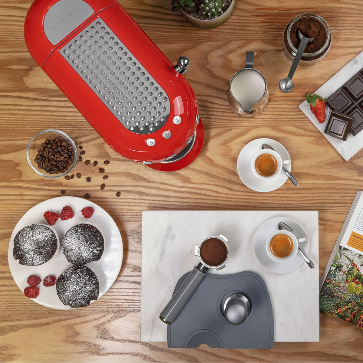 Draufsicht auf eine rote Kaffeemaschine, Kaffeetassen, Schokolade, Muffins, Kaffeebohnen und eine Zeitschrift auf einem Holztisch.