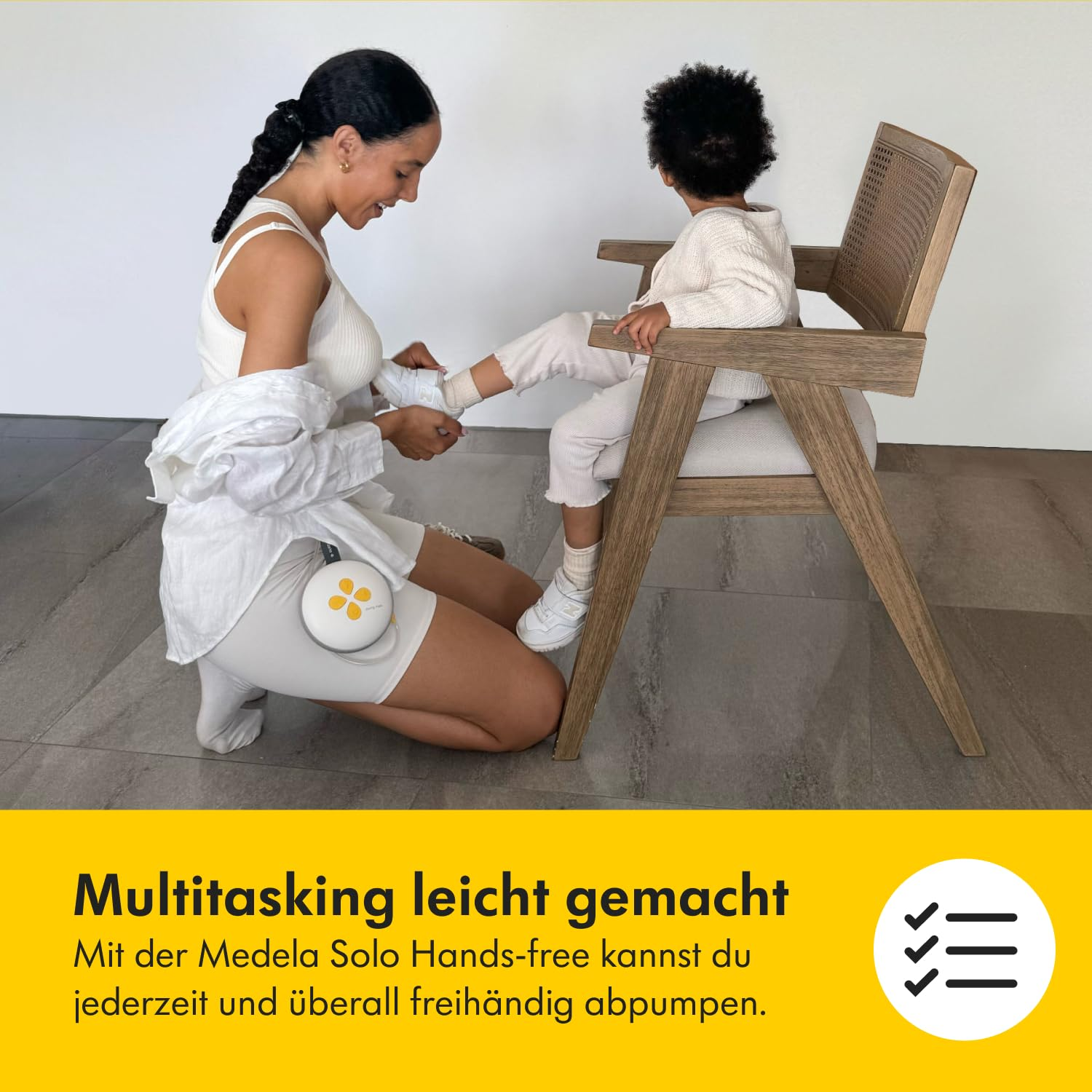 Eine Frau hilft einem Kind beim Anziehen der Schuhe. In der Nähe steht eine Brustpumpe. Das Kind sitzt auf einem Holzstuhl.